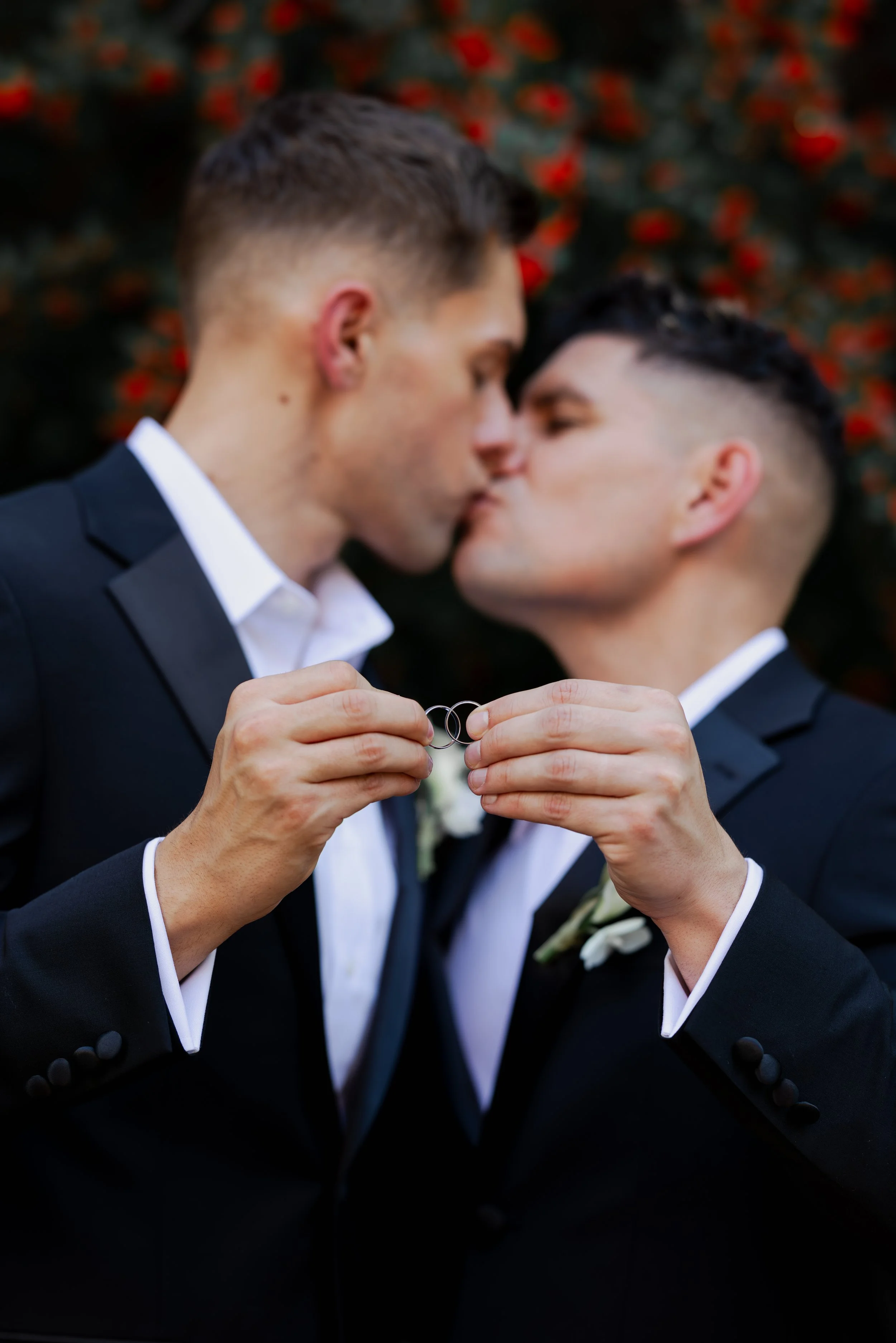 Two men in black tuxedos kiss while holding wedding rings in front of a blurred background of red leaves.