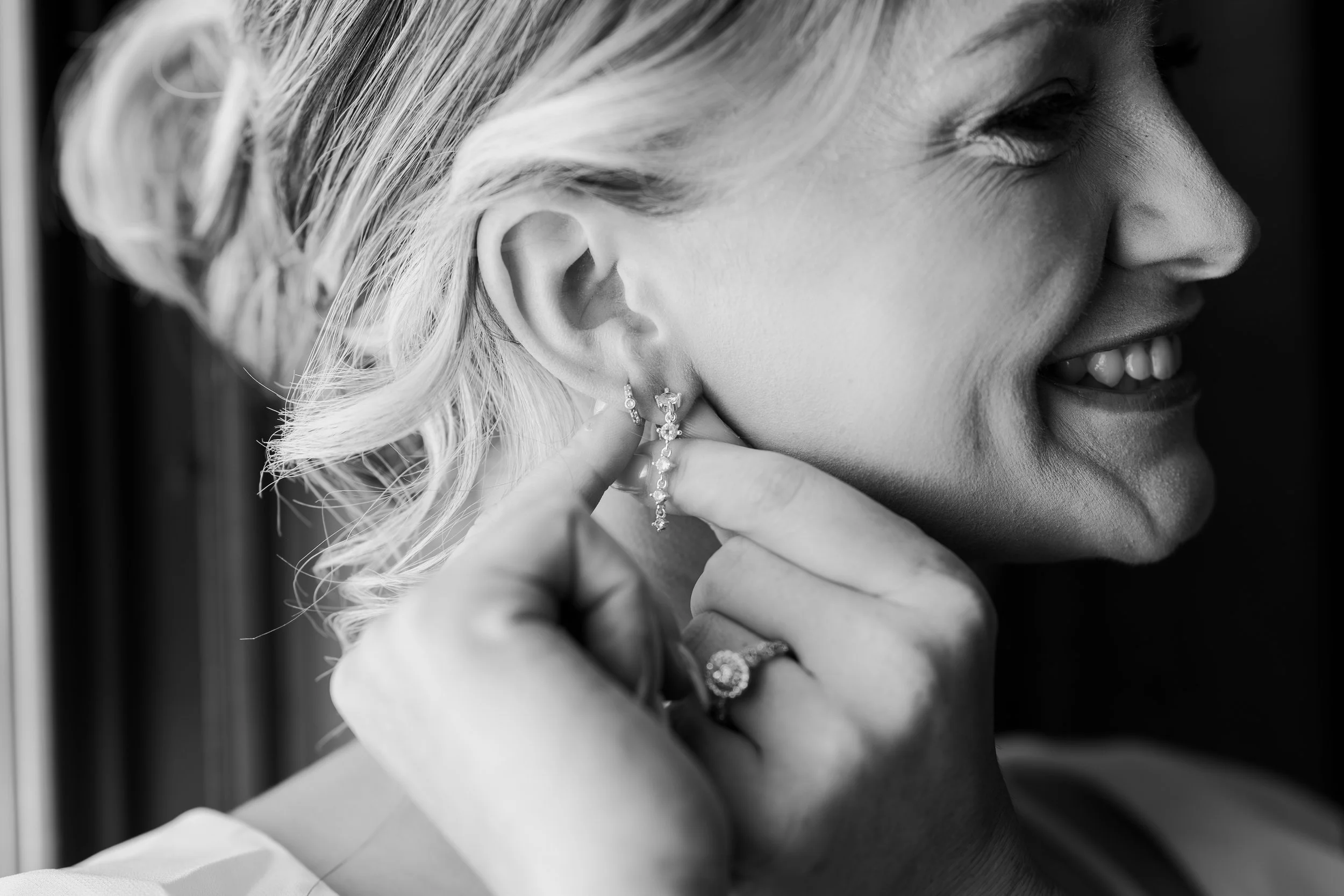 Close-up black and white photo of a woman smiling as she puts on earrings, with a wedding ring visible on her finger.