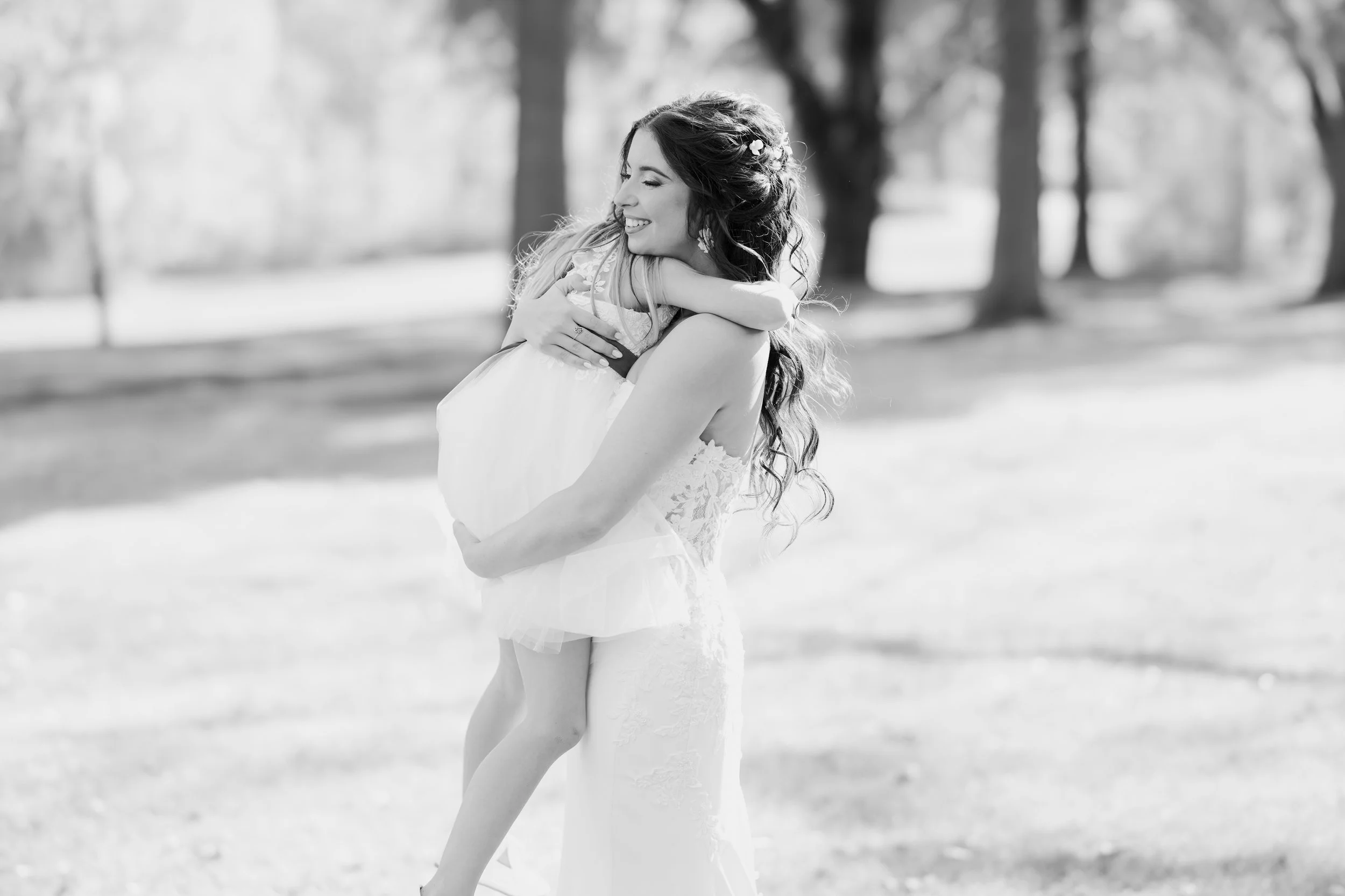 A woman in a wedding dress embraces a young girl in a park, both smiling. The woman has long, wavy hair and is wearing a lace wedding gown. The girl is wearing a light-colored dress with a tulle skirt. In the background, there are trees and an outdoo