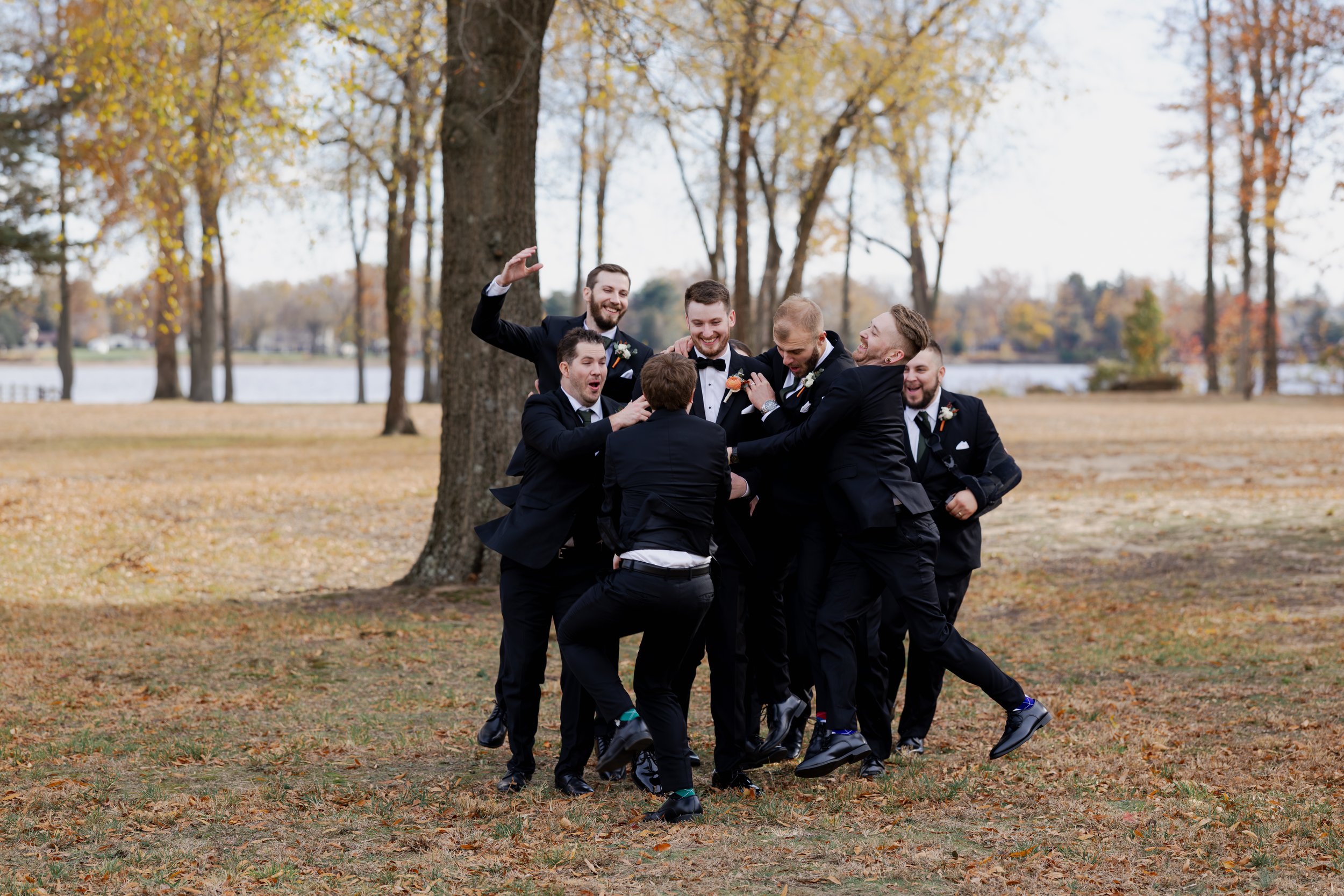 Group of men in suits celebrating outdoors in fall, with trees and lake in the background.