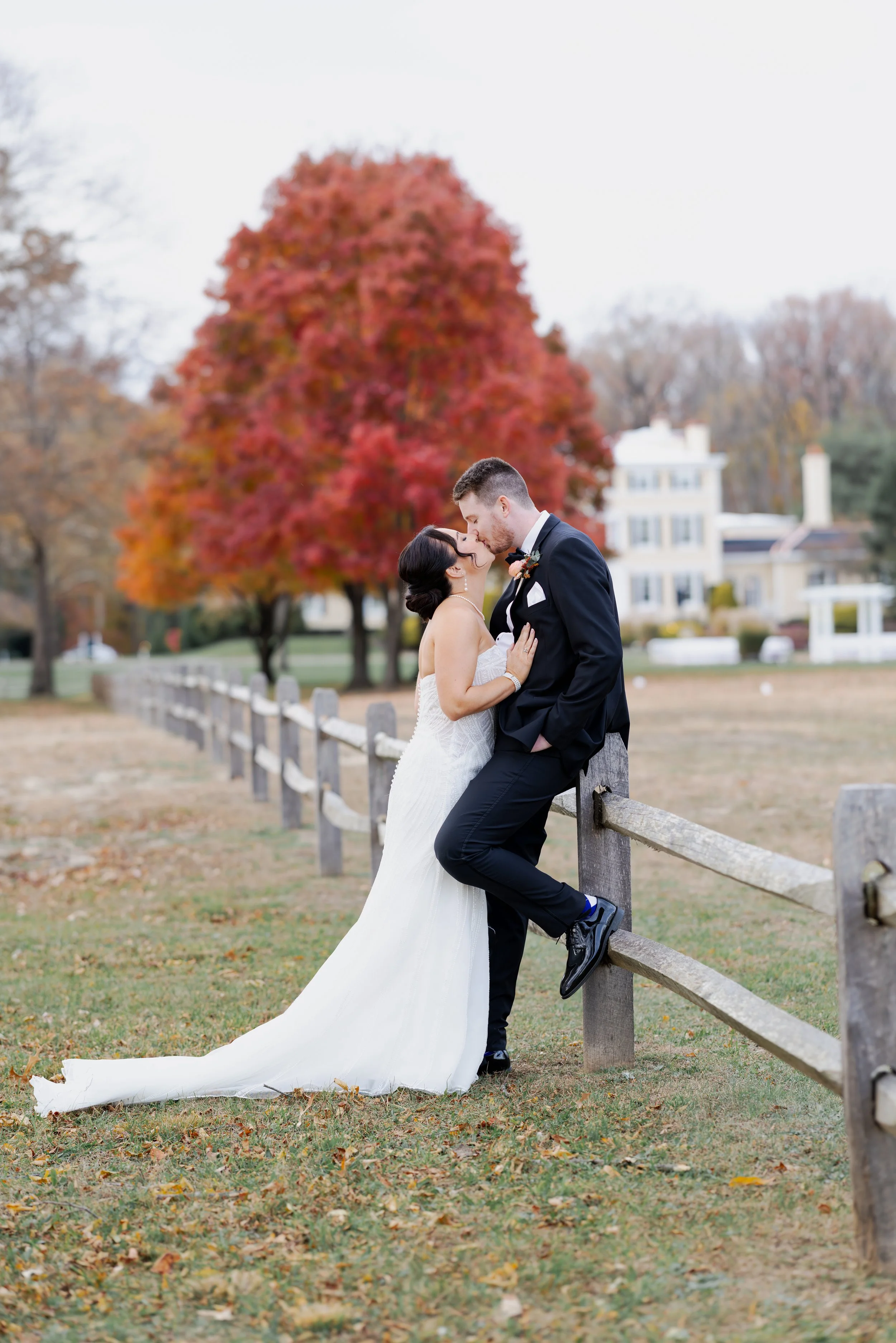 A newlywed couple sharing a kiss outdoors on a fence, with a large red tree and a white house in the background during fall.