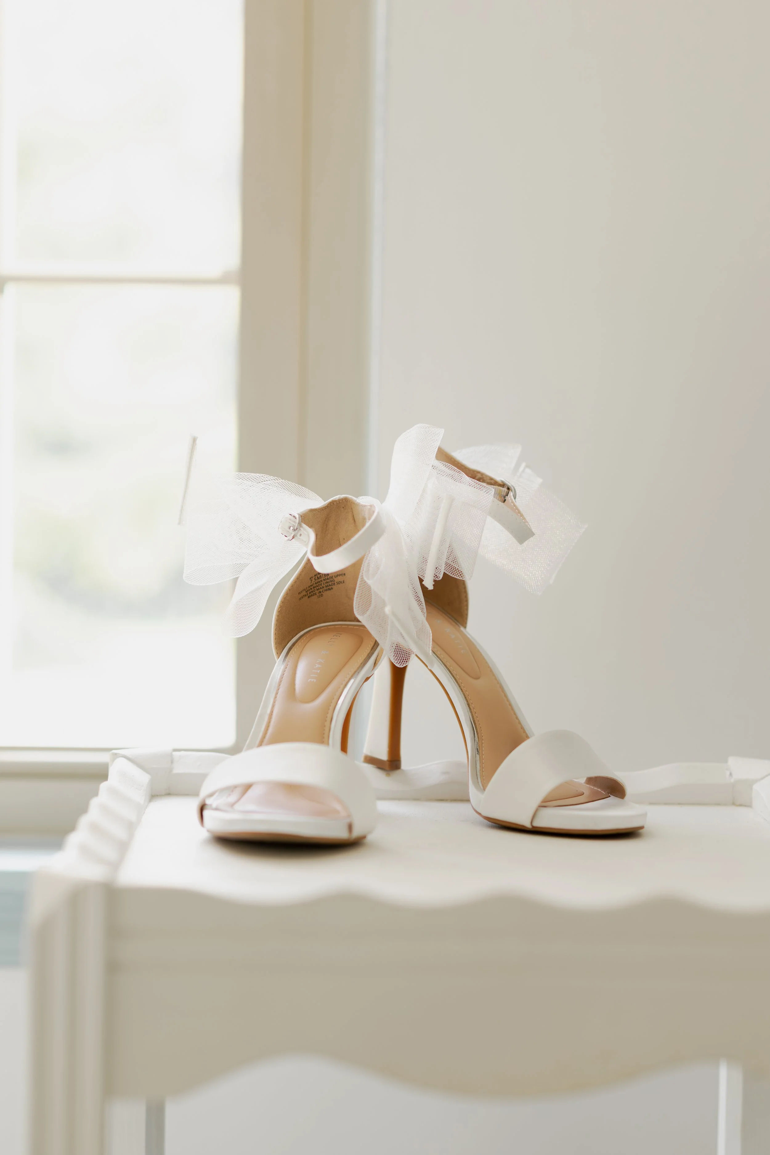 Close-up of white high-heeled shoes with ankle straps and tulle bows, placed on a white surface near a window.