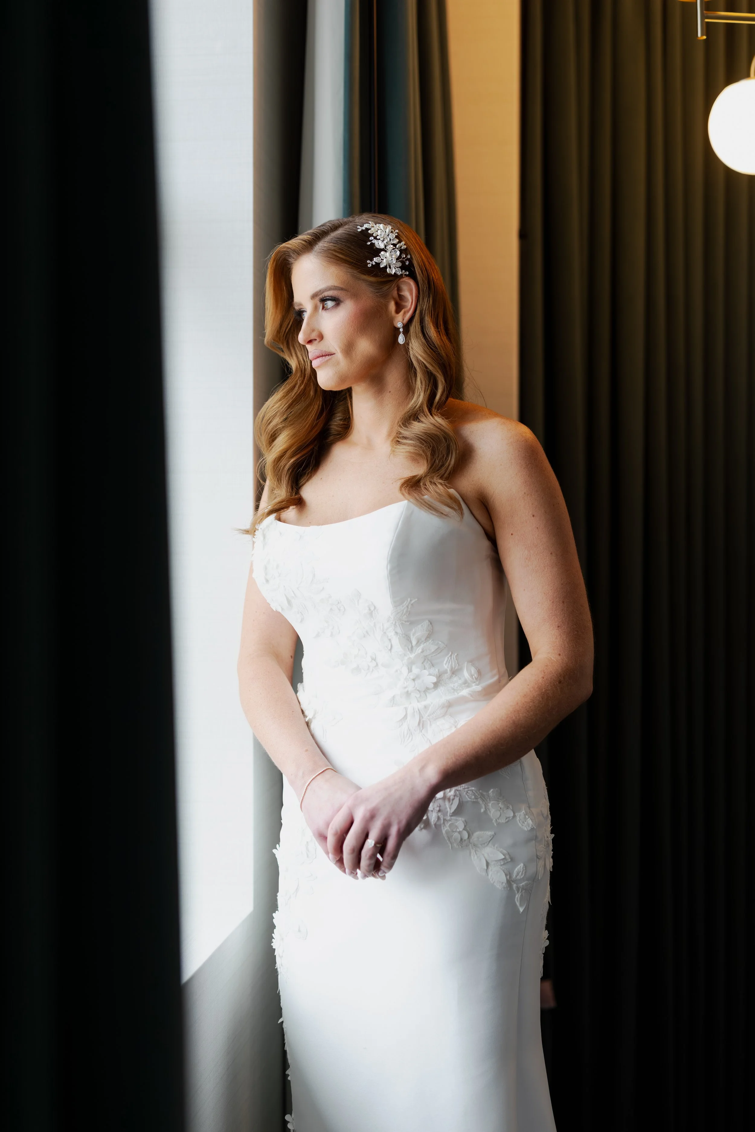Bridal woman standing by a window, looking outside, wearing a white wedding gown with floral embroidery, earrings, and a floral hair accessory, with dark curtains and warm lighting in the background.
