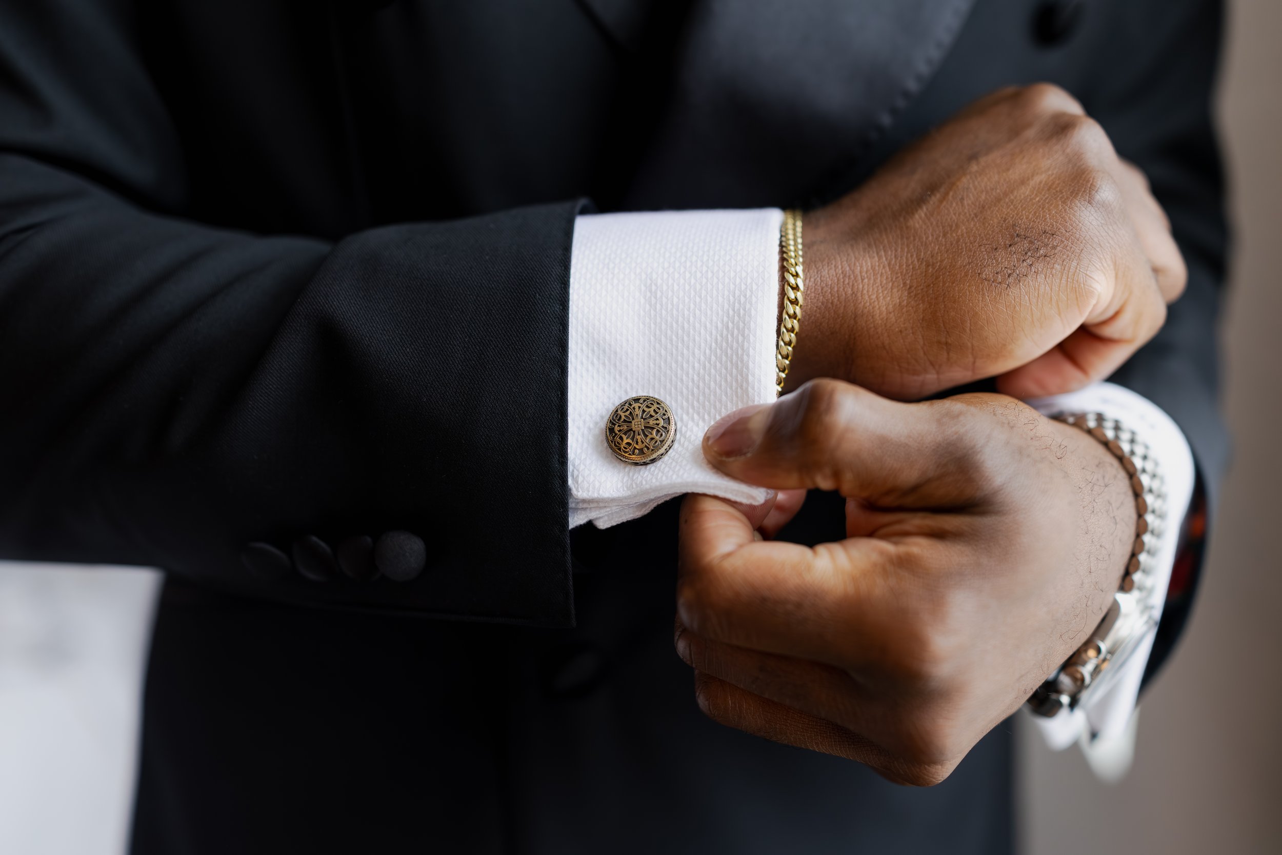 A man adjusting the cuff of his black suit jacket, revealing white shirt cuffs with silver cufflinks, a gold bracelet, and a silver watch.