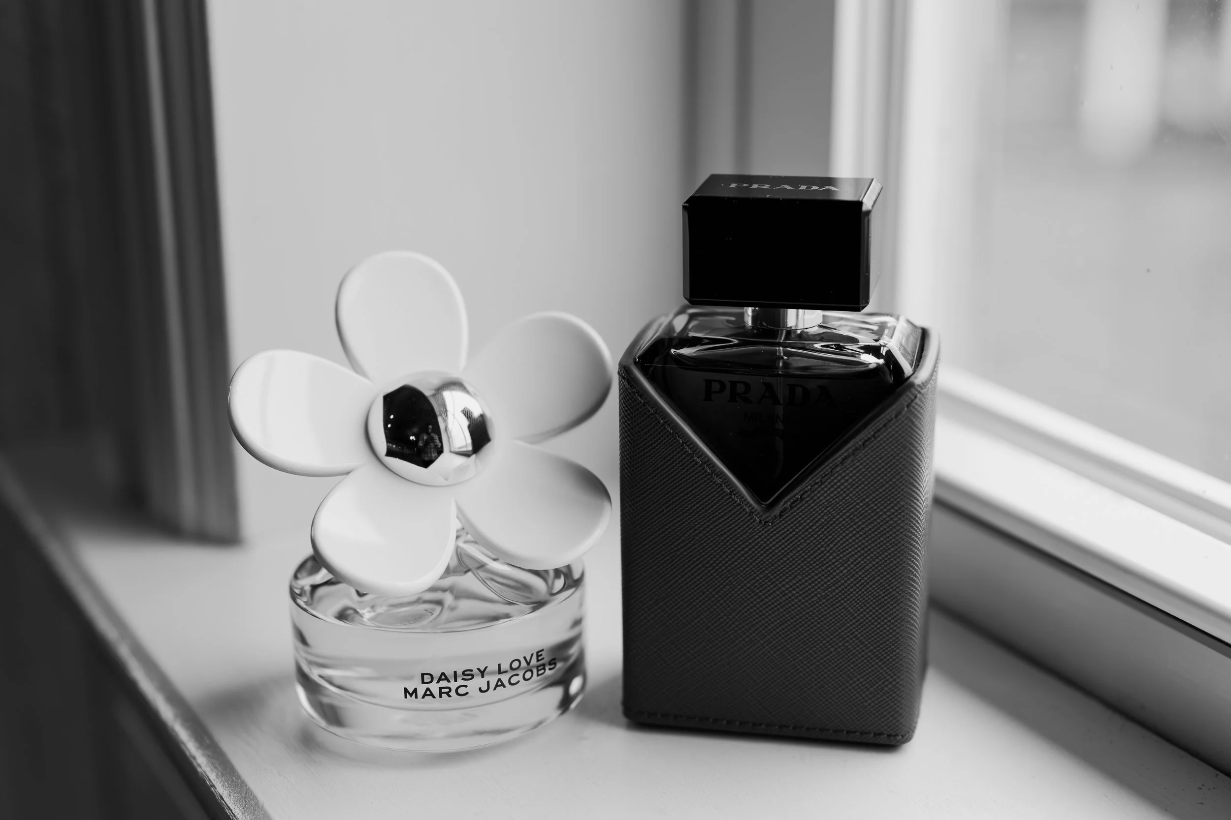 Black and white photo of two perfume bottles on a windowsill, with a flower-shaped decoration on one. One bottle is labeled 'Daisy Love Marc Jacobs' and the other has a Prada logo.