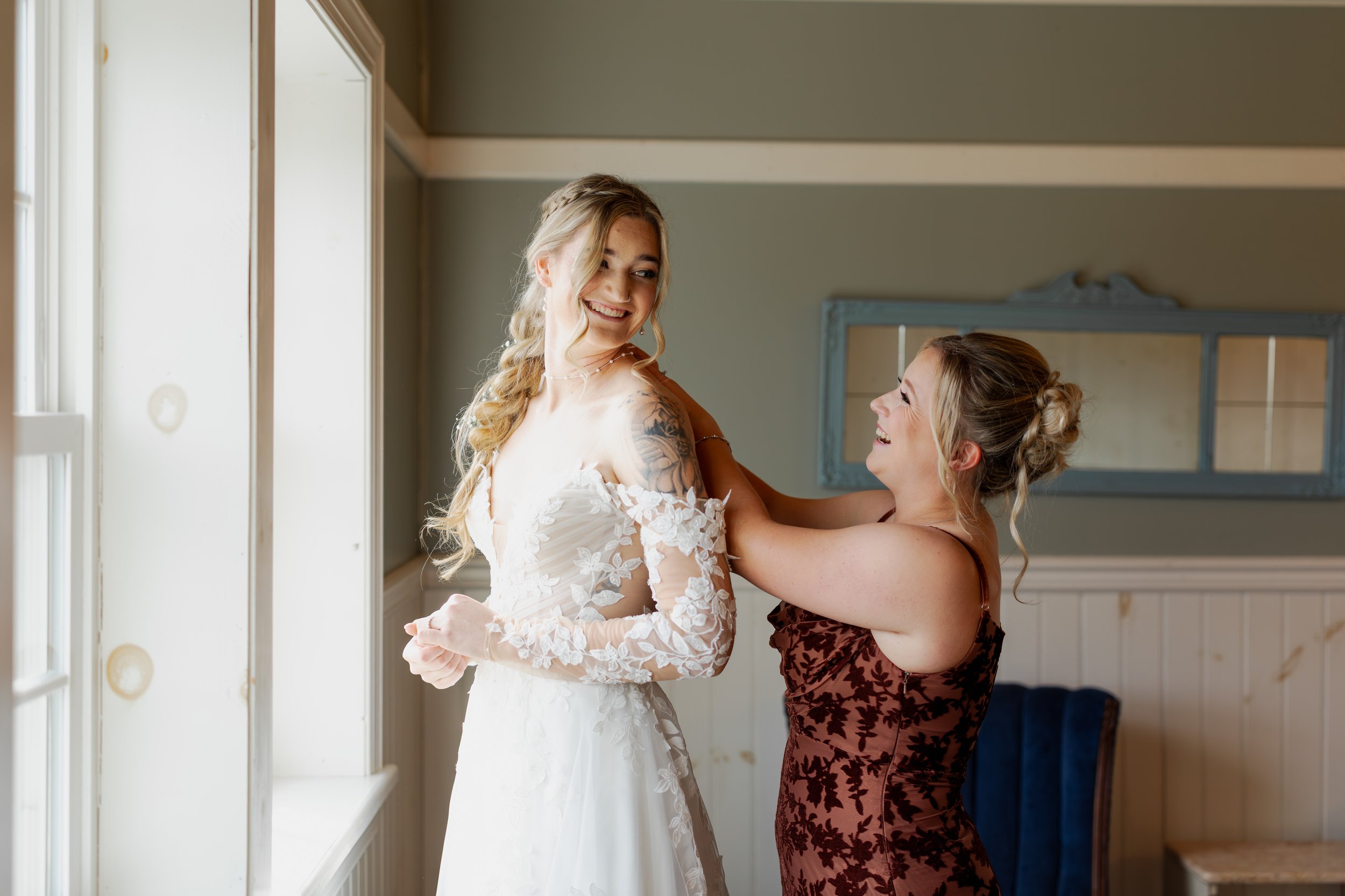 Two women, one in a white wedding dress and the other in a burgundy dress, smiling and preparing for a wedding in a well-lit room.