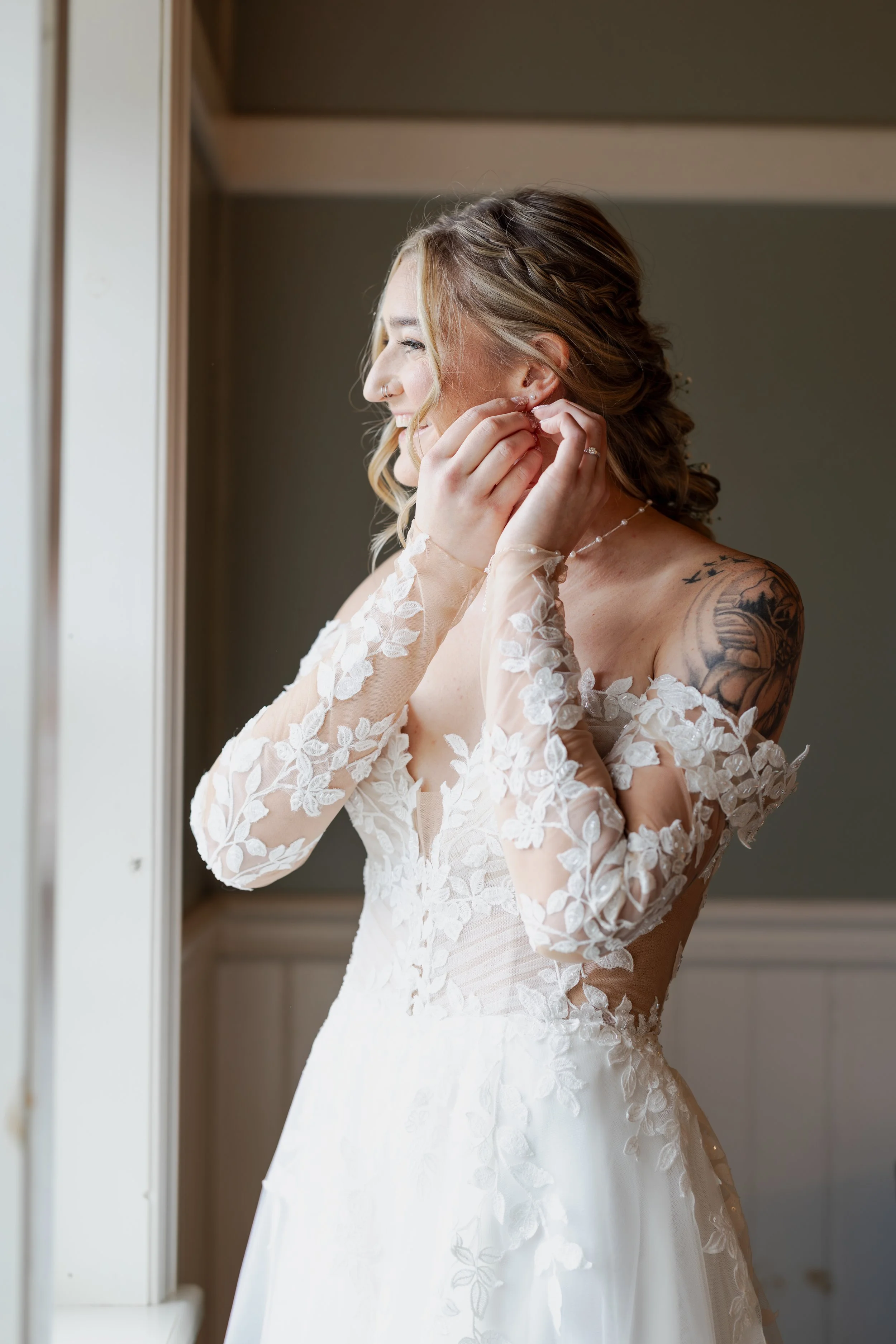 A woman in a white wedding dress with lace sleeves is putting on earrings by a window, smiling, with tattoos visible on her shoulder.