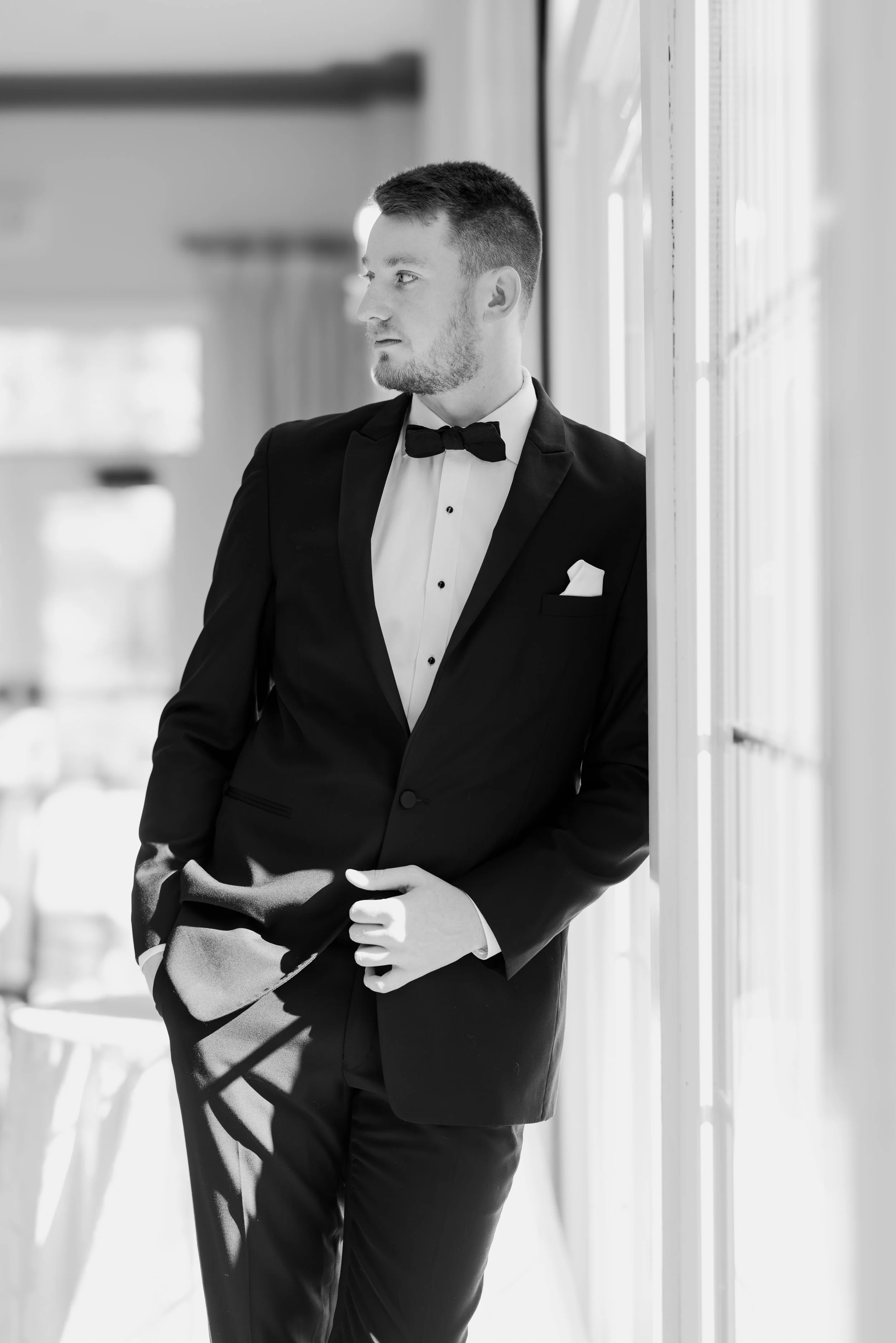 A man in a tuxedo with a bow tie, standing near a window, holding a coat in his hand, looking thoughtfully outside.