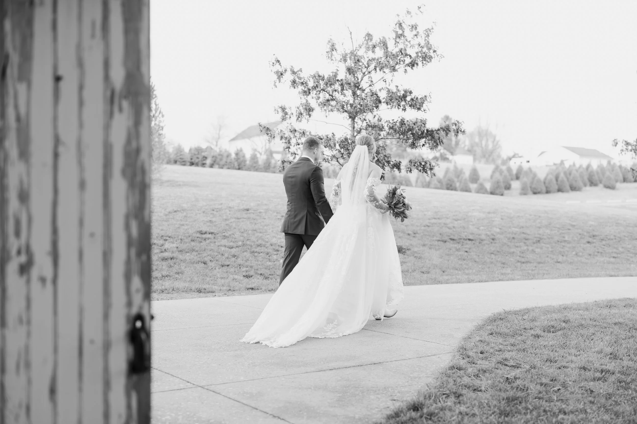 A bride and groom walking together outdoors on a sidewalk, with the bride holding a bouquet and wearing a long wedding dress and veil, seen from behind. They are near a large tree on a grassy area with houses in the background.