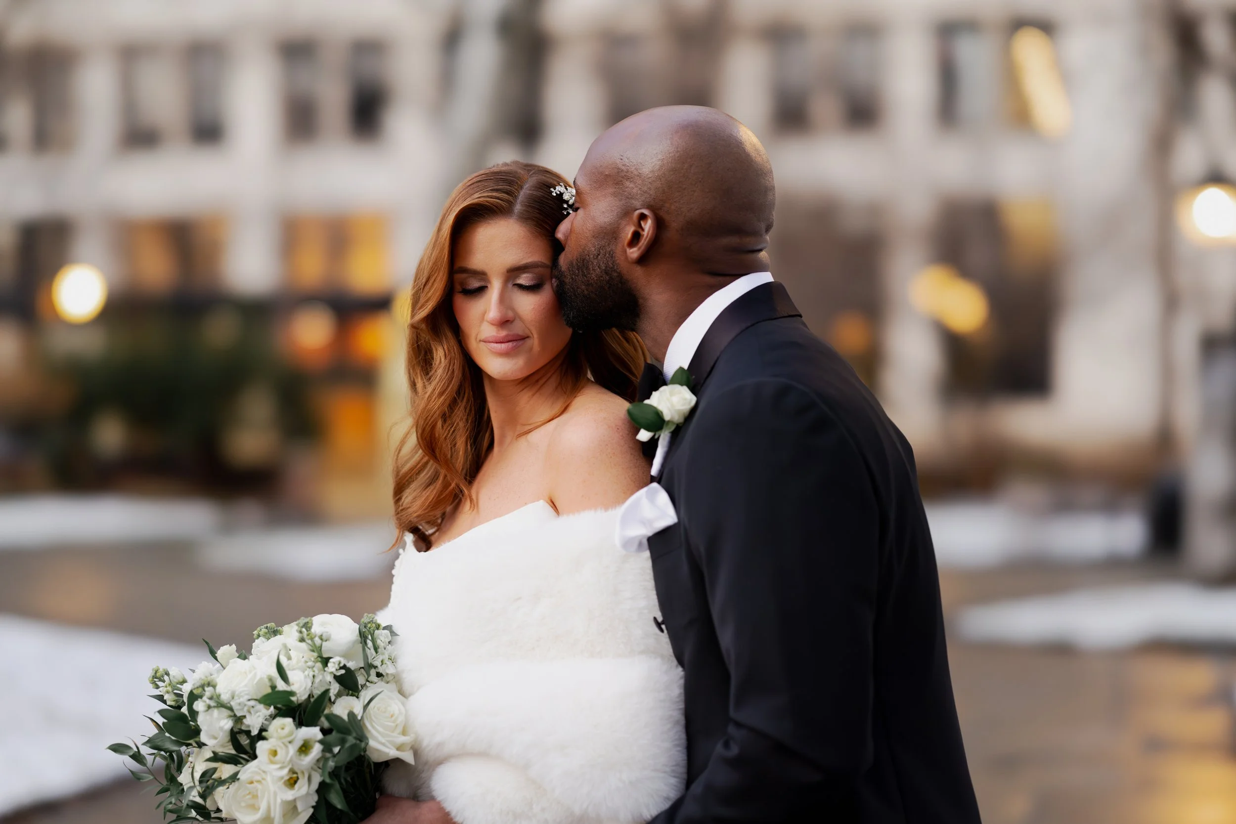 A bride and groom embrace outdoors in winter, with the groom kissing the bride's forehead; she holds a bouquet of white roses and greenery.