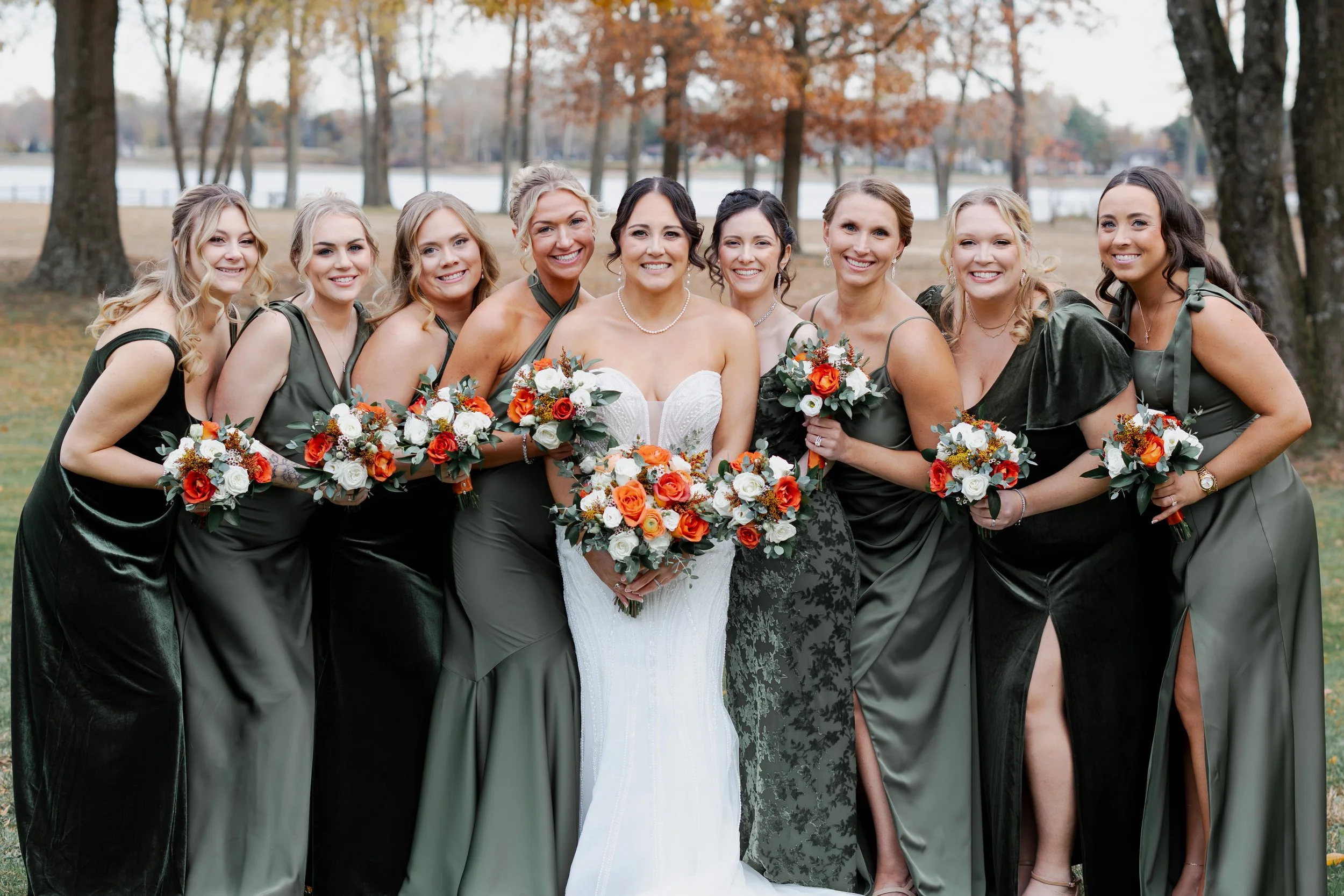 Bride in white wedding gown surrounded by bridesmaids in dark green dresses holding bouquets outdoors near a lake and trees with autumn leaves.