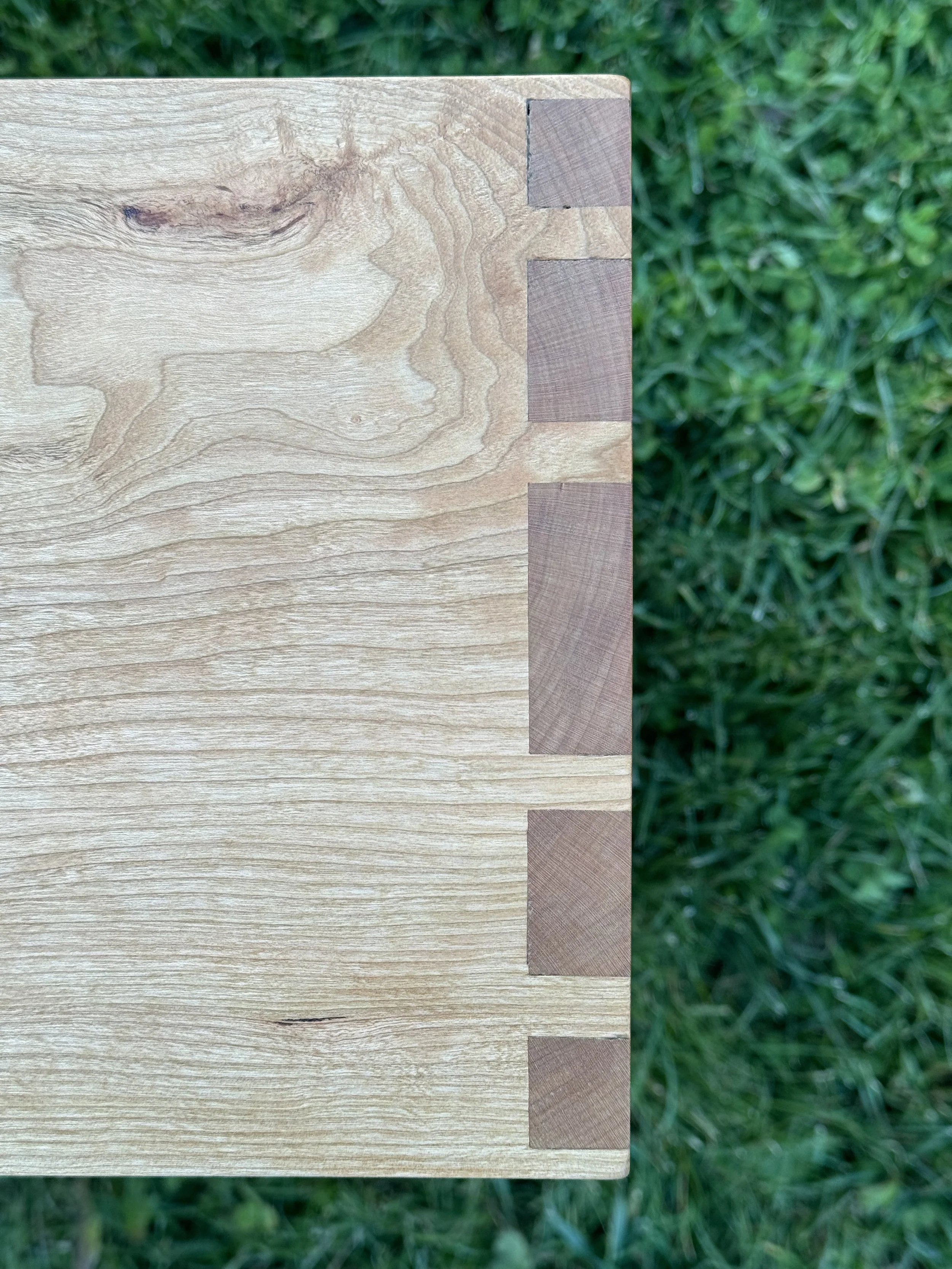 Close-up of a wooden table corner with dovetail joints, outdoors on green grass.