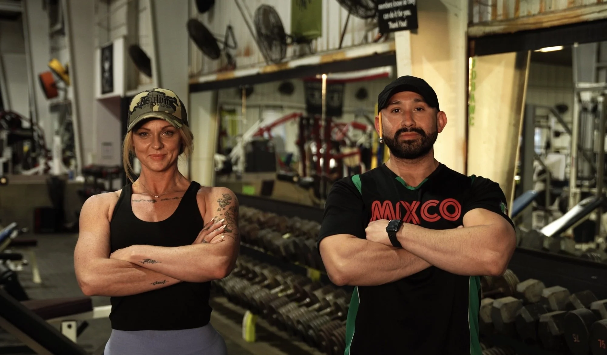 Two fit individuals, a woman with tattoos and a woman with a beard, standing in a gym with arms crossed.