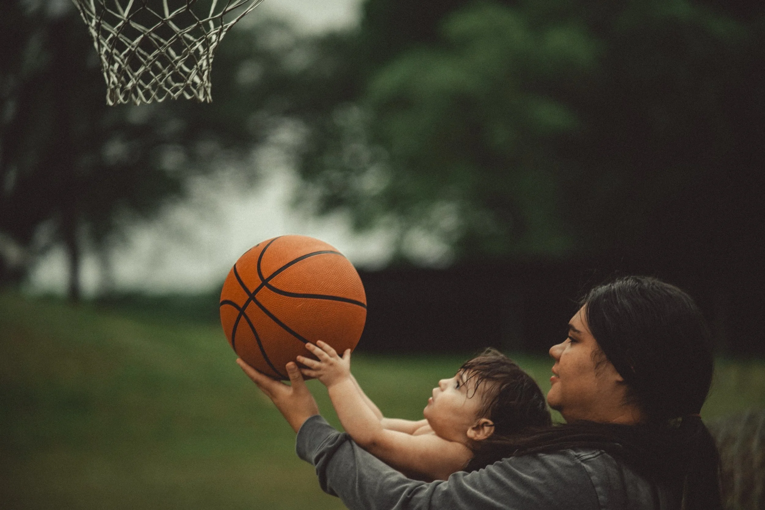 A woman holding a young child as they prepare to shoot a basketball towards a hoop outdoors on a cloudy day.