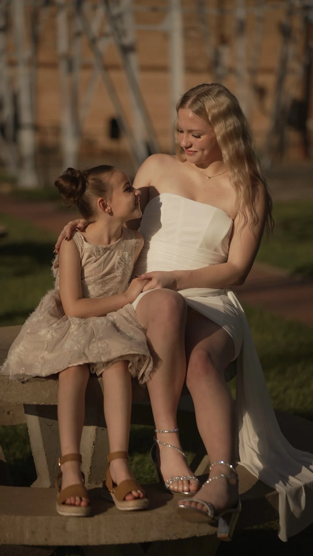 A woman in a white strapless dress and a young girl in a beige dress with a floral pattern sitting close together on a bench, sharing a tender moment outdoors during sunset.