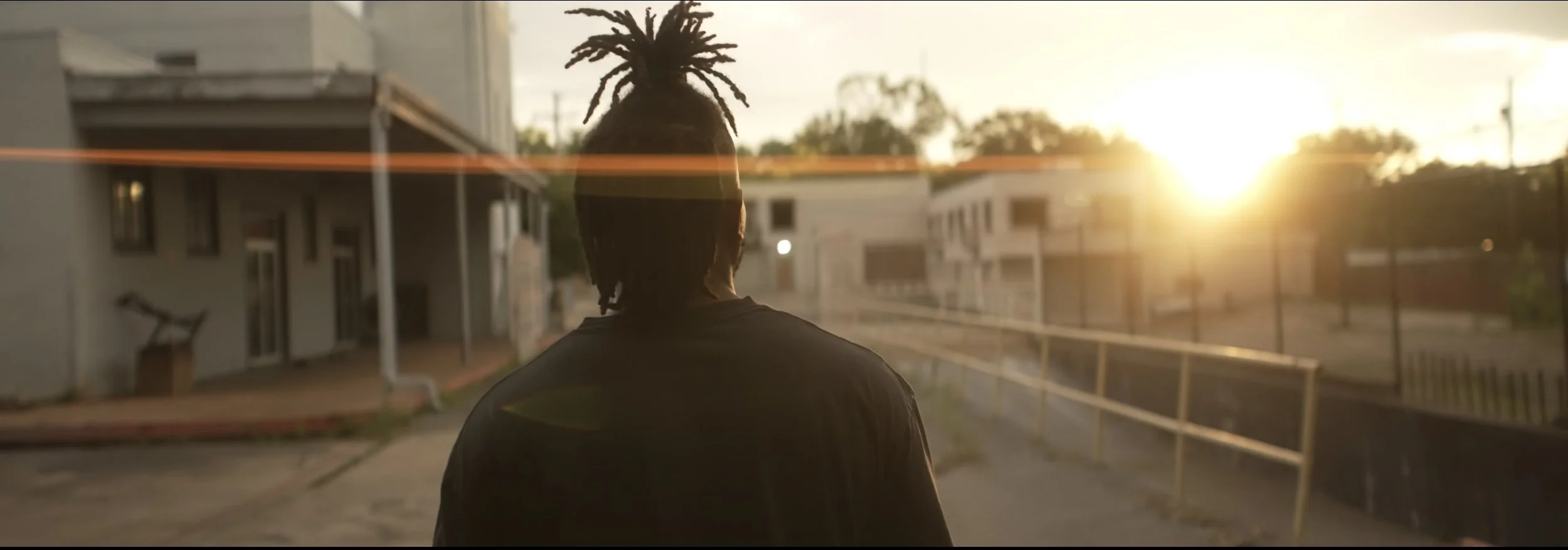 A person with dreadlocks standing outdoors during sunset, facing away from the camera, with residential buildings and a playground area in the background.