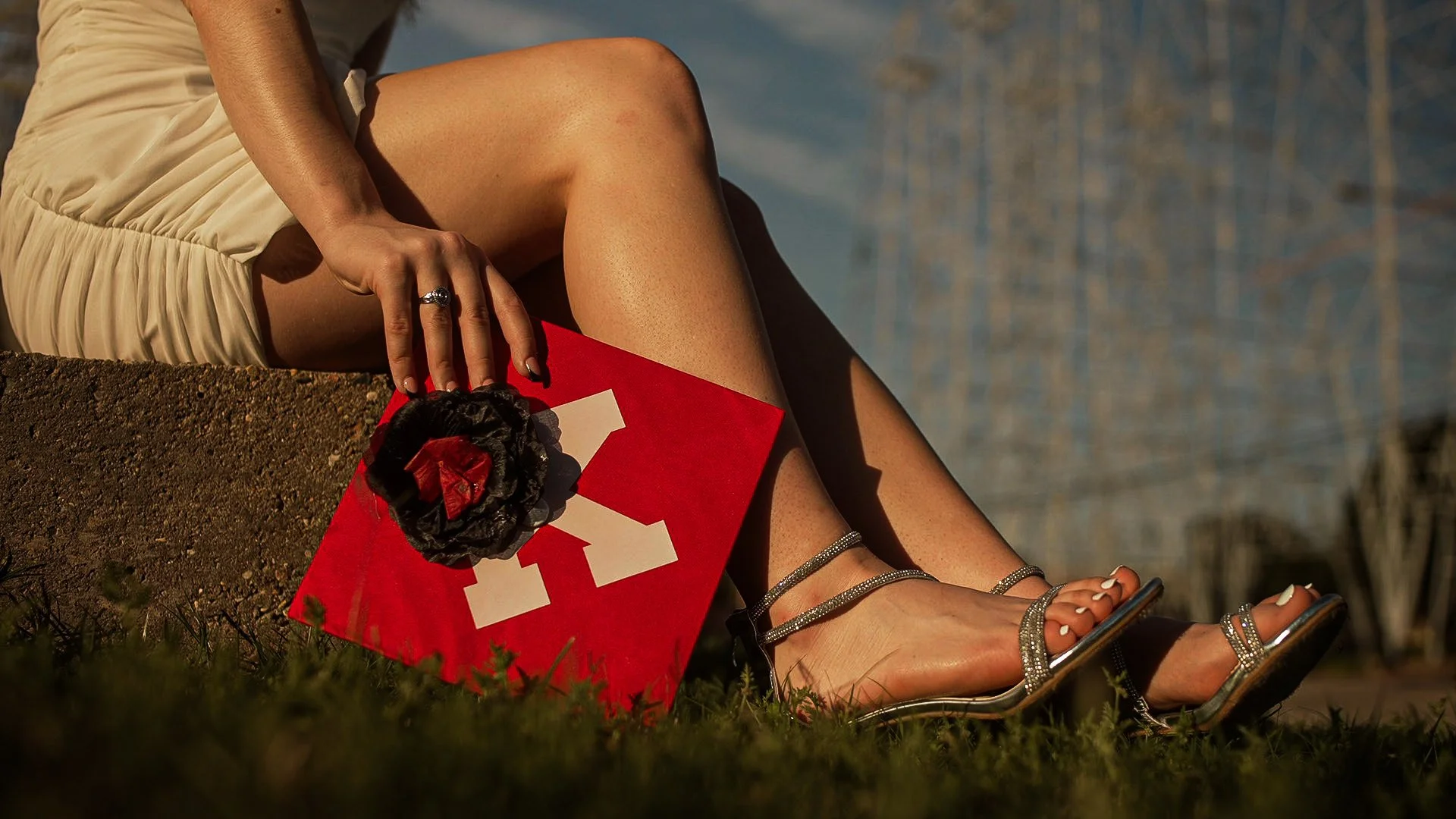 Close-up of a woman sitting outdoors on grass, wearing beige dress and silver high-heeled sandals, holding a red book with a black flower and white ribbon decoration, near a body of water.