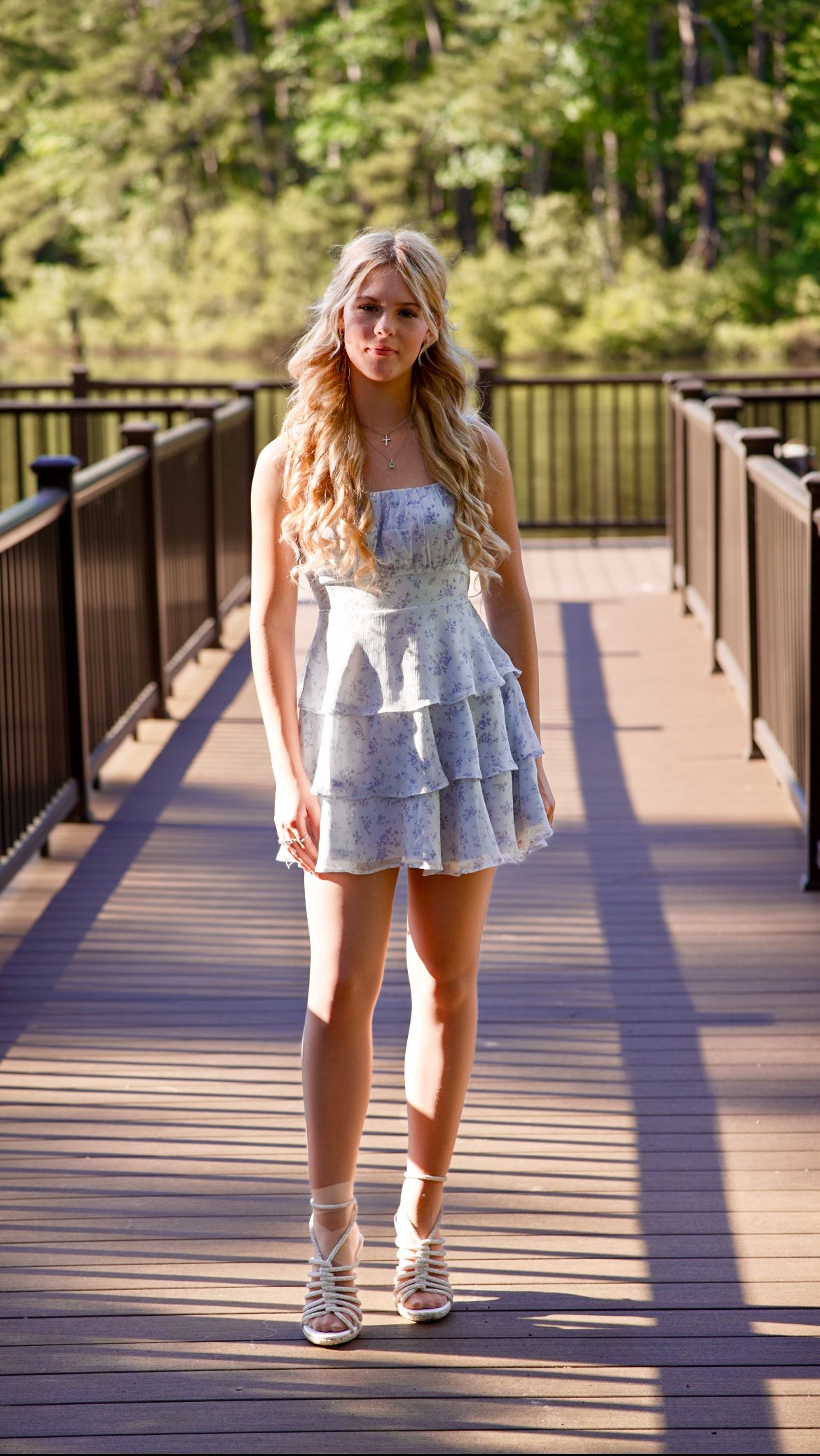 A young woman with long blonde hair standing on a wooden bridge surrounded by green trees, wearing a light-colored floral dress and strappy heels.