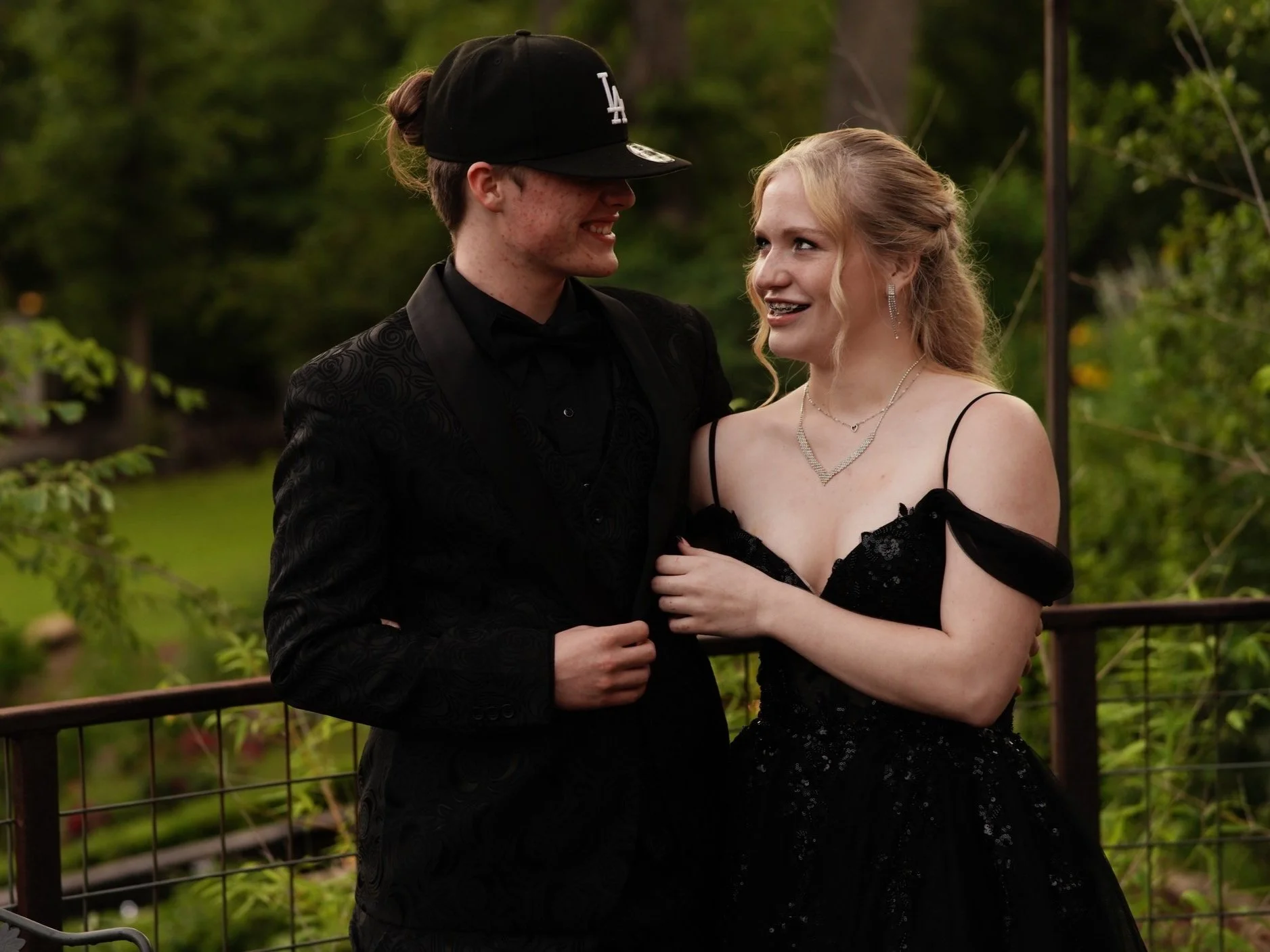 A young man and woman dressed in formal attire smiling and looking at each other outdoors, with greenery in the background.