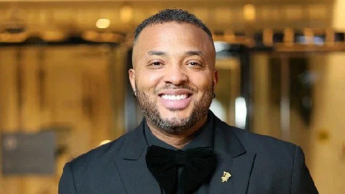 A smiling man dressed in a black tuxedo with a bow tie, standing indoors with a blurred background.