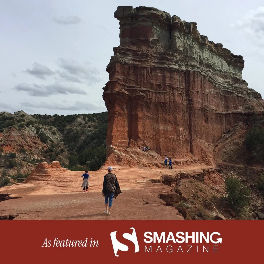 A group of people walking near a large, layered red sandstone rock formation in a desert-like landscape.