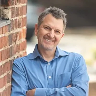 Smiling man with gray hair and beard, wearing a blue button-up shirt, leaning against a brick wall outdoors.