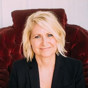 A woman with blonde hair and a black blazer sitting on a red upholstered armchair, smiling at the camera.