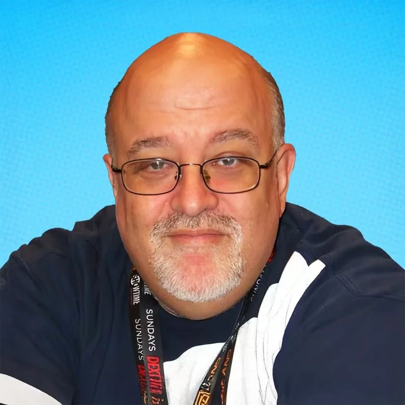 A middle-aged man with glasses, a short beard, and a bald head, wearing a black t-shirt with white stripes, smiling against a bright blue background.