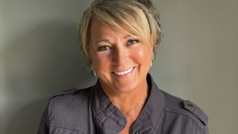 A woman with short blonde hair smiling, wearing a gray military-style jacket, standing against a plain gray background.