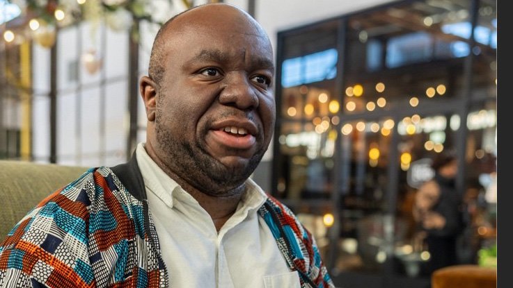 A man with dark skin, wearing a patterned shirt over a white collared shirt, sitting indoors at a restaurant or café with blurred warm lights and people in the background.