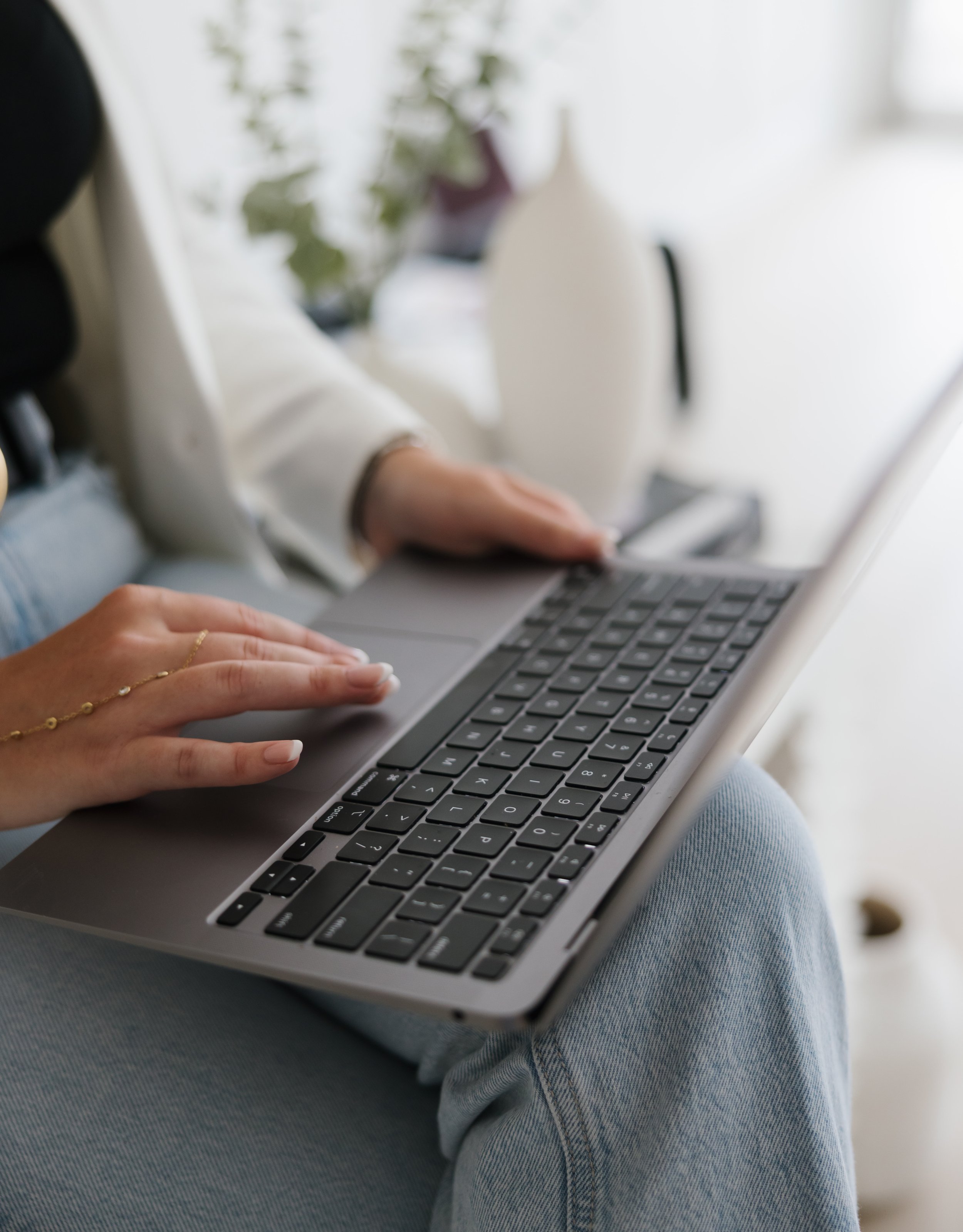 Person using a laptop on their lap, sitting on a chair or couch with a blurred background of plants and decor.