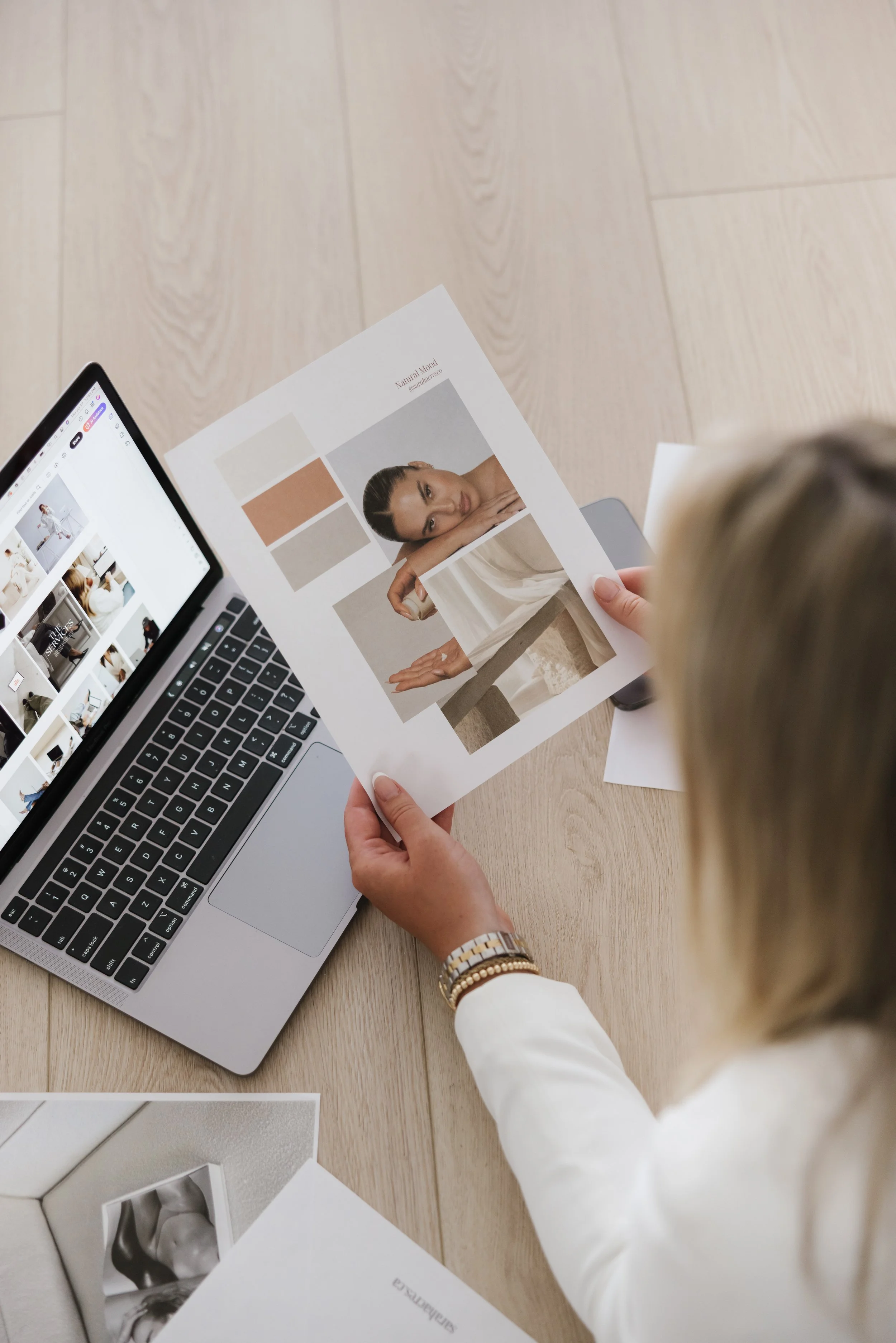 A woman looking at a photo sheet with a laptop open next to her on a wooden table.