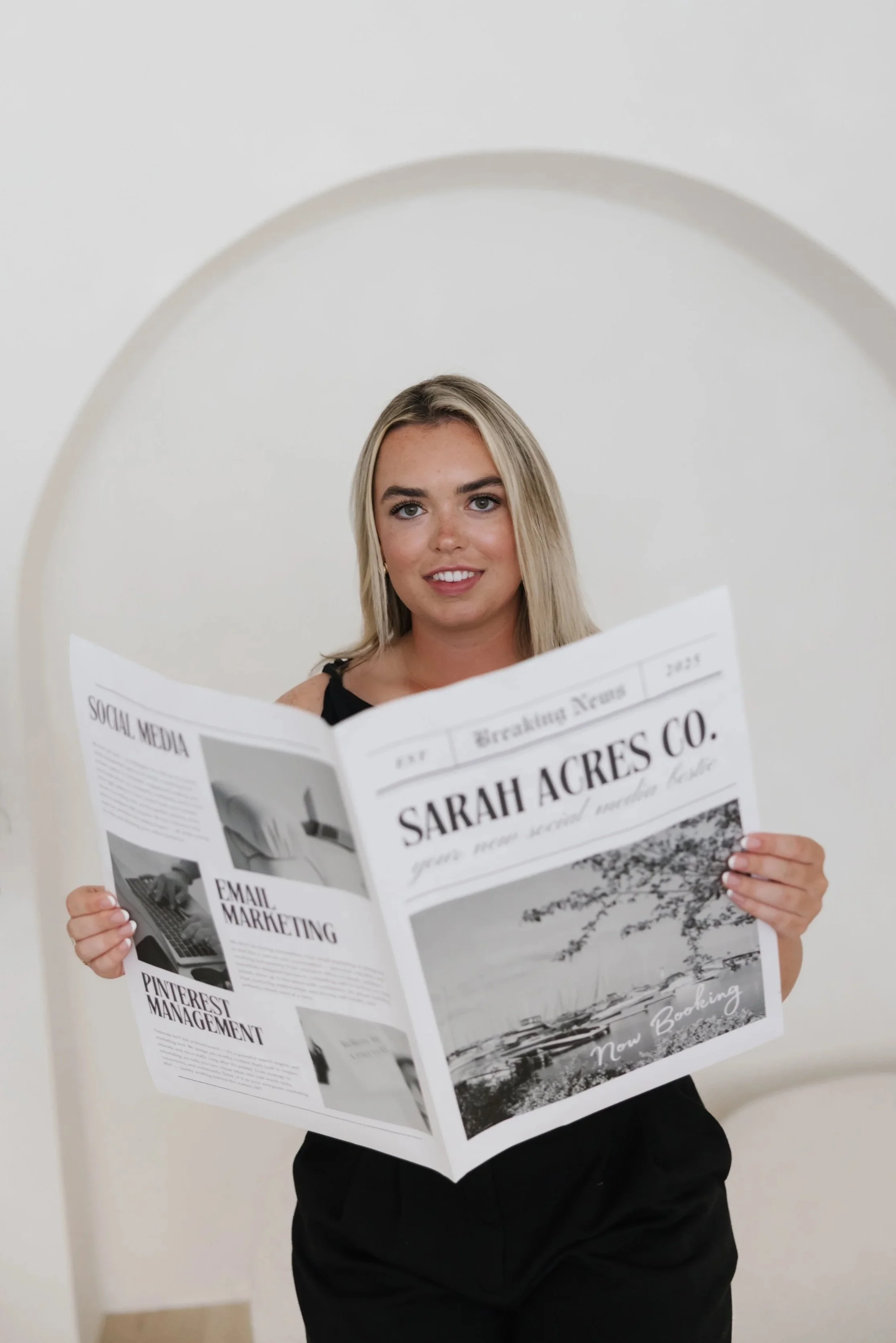 A woman with blonde hair and a black sleeveless top stands in front of a white wall, holding a newspaper titled 'Sarah Acres Co' with various articles and images, smiling at the camera.