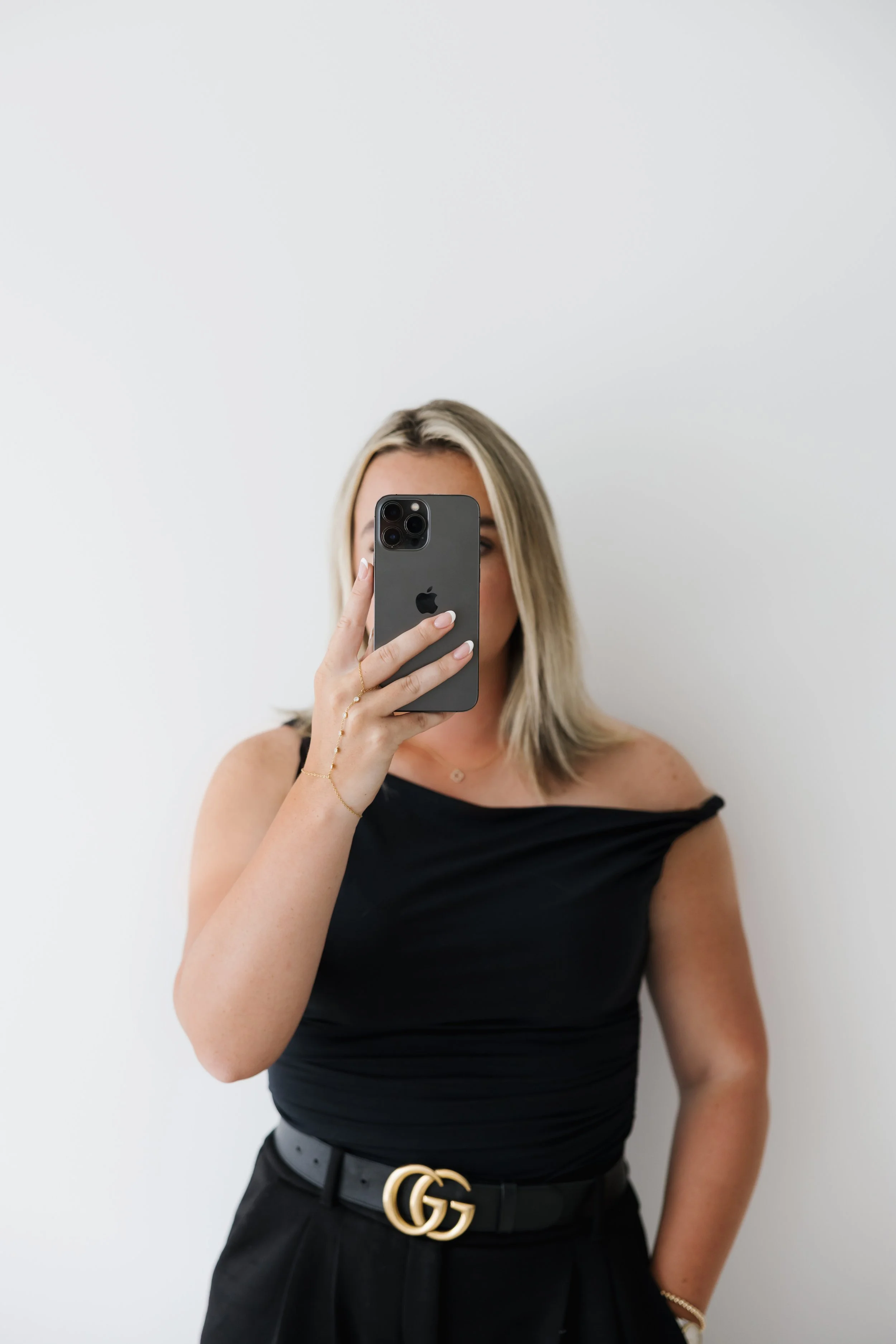 Woman with blonde hair wearing a black off-the-shoulder top and black belt with gold GG buckle taking a selfie with an iPhone in front of a plain white wall.