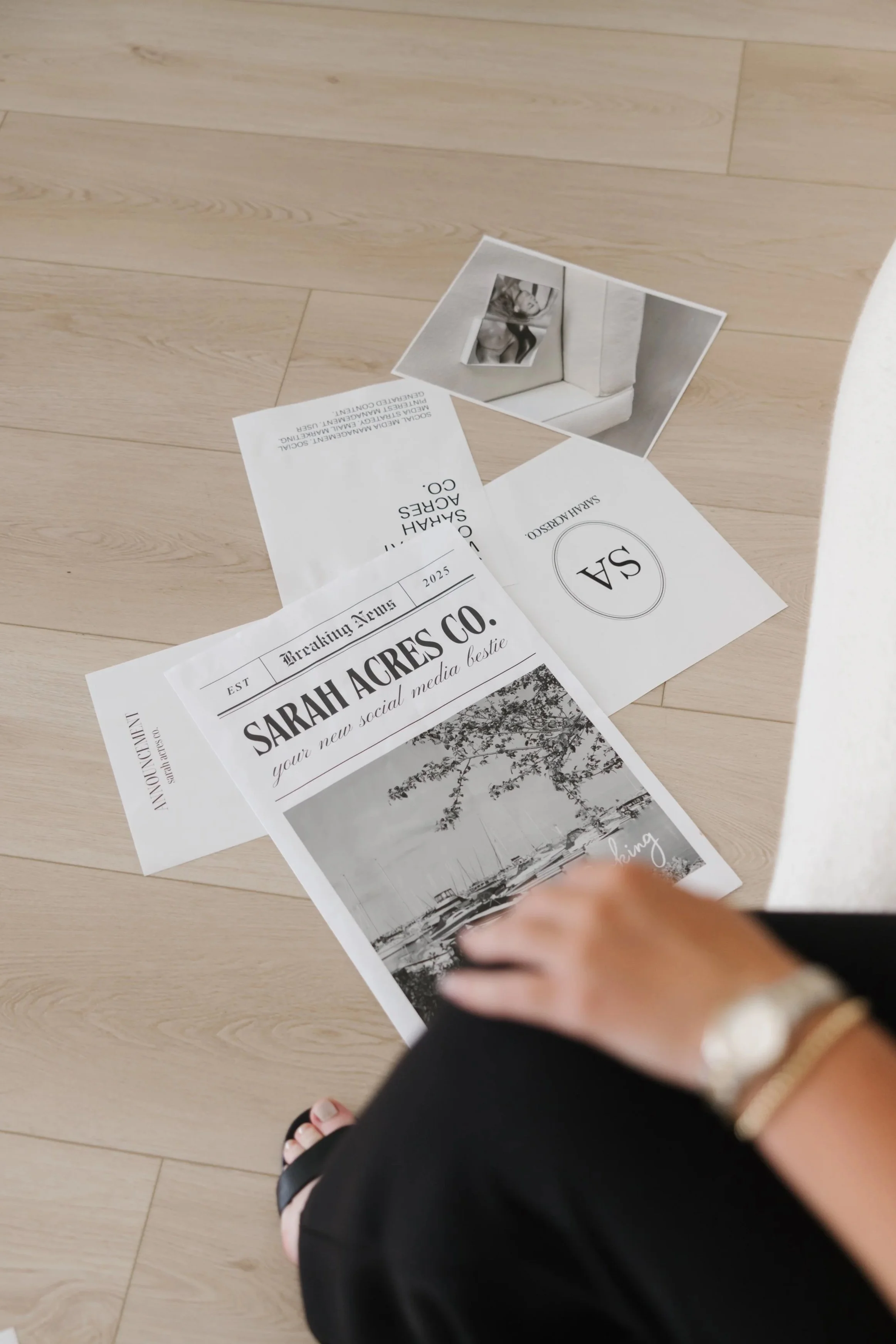 Various papers and photographs scattered on a wooden floor, including a black and white image of a boat, a social media company flyer, and a magazine page, with part of a person's hand and foot visible in the foreground.