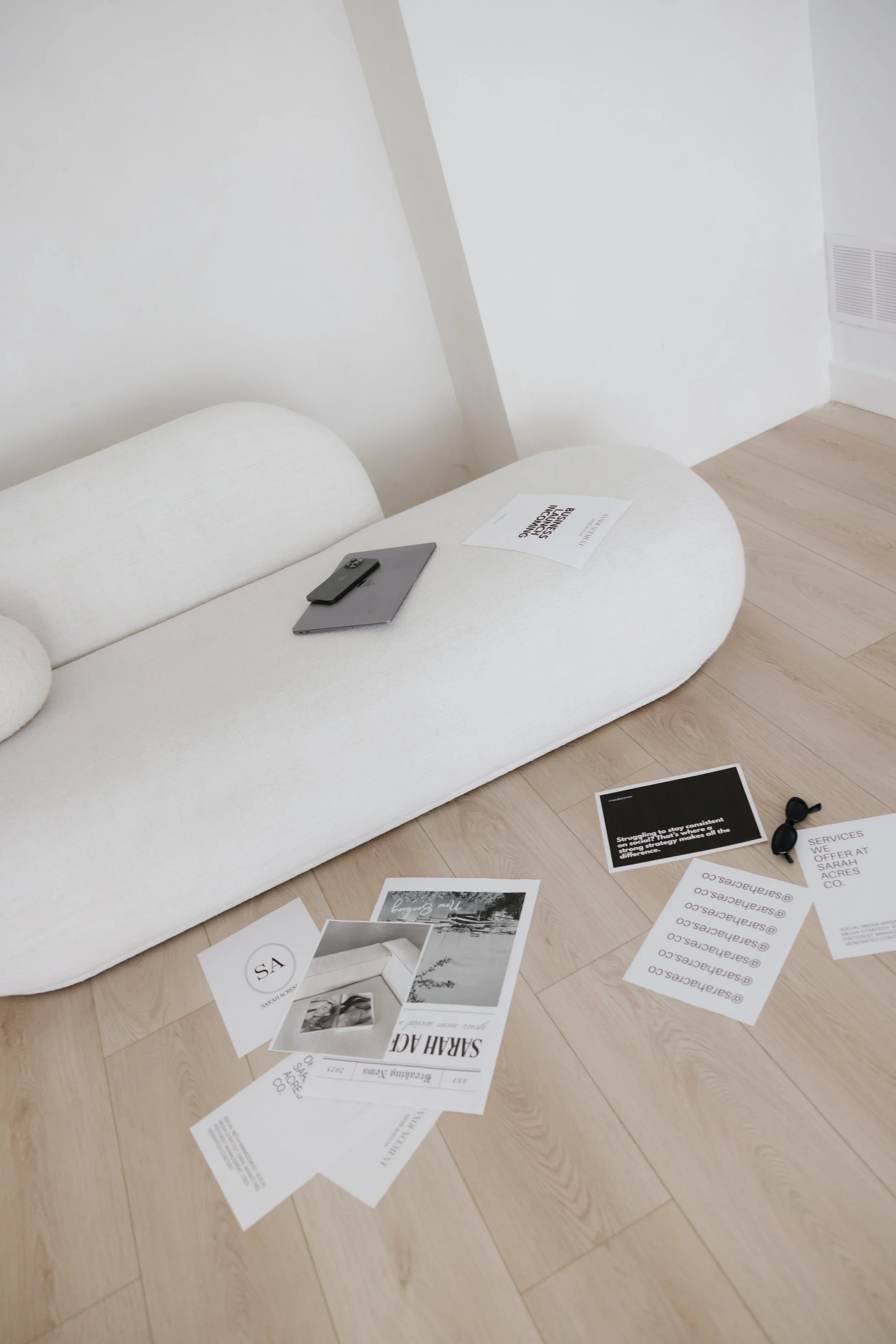 White sofa with various printed papers and a mobile phone on it, scattered papers on the wooden floor next to the sofa, in a minimalist room.