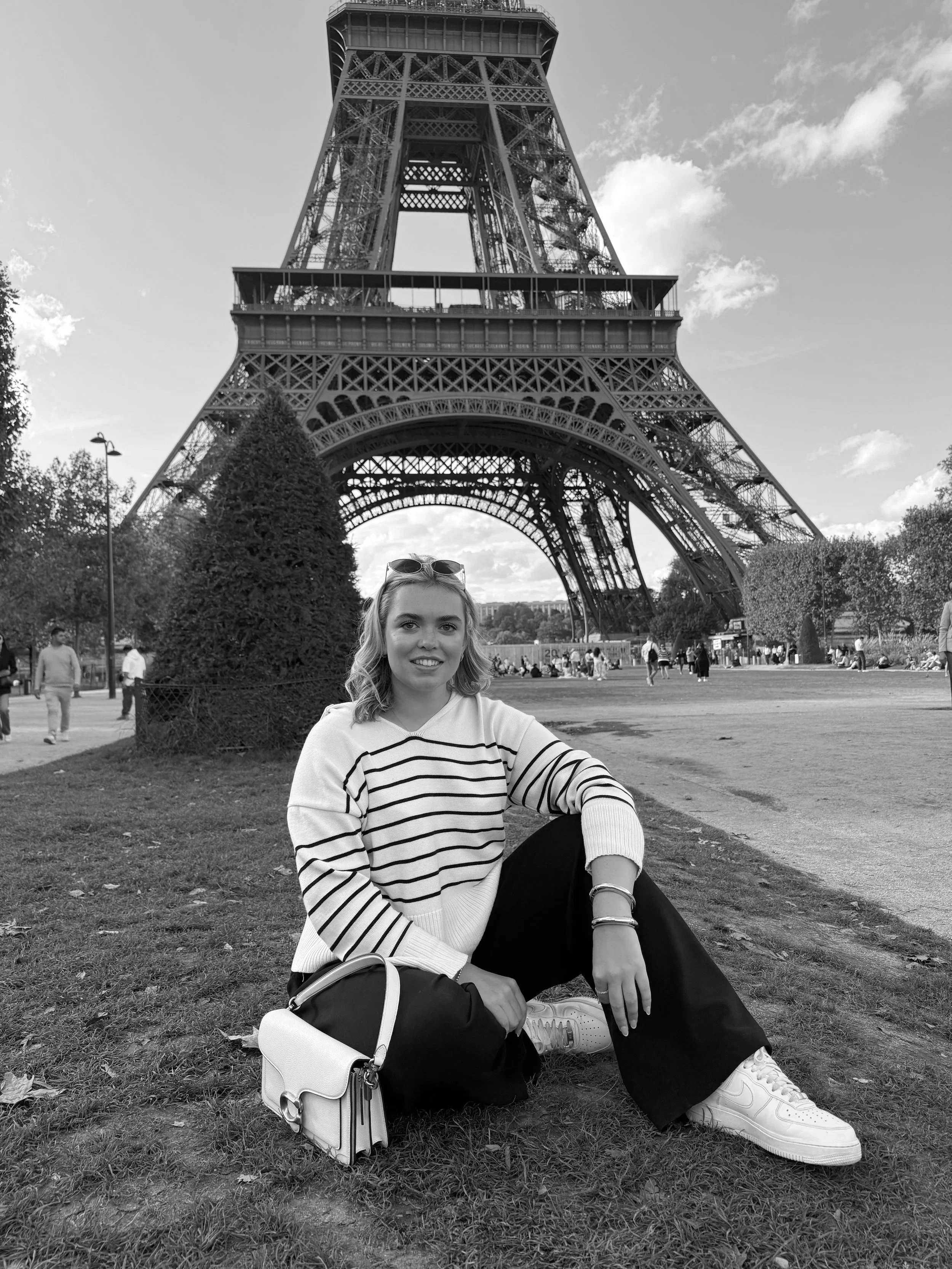 A woman sitting on the grass in front of the Eiffel Tower in Paris, France, with trees and other visitors in the background.