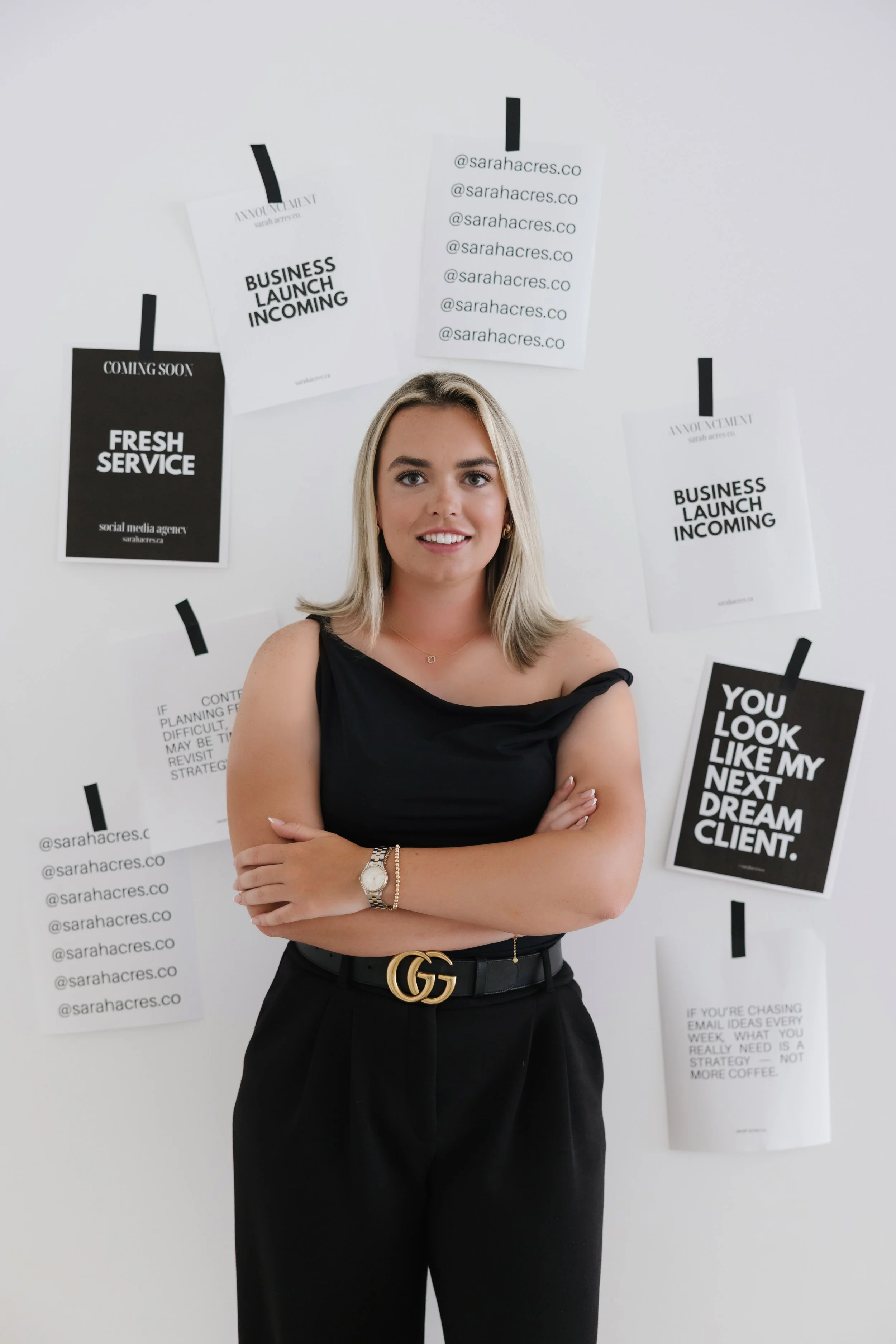 A young woman with shoulder-length blonde hair stands with crossed arms in front of a white wall decorated with black and white posters and notes about a business launch.