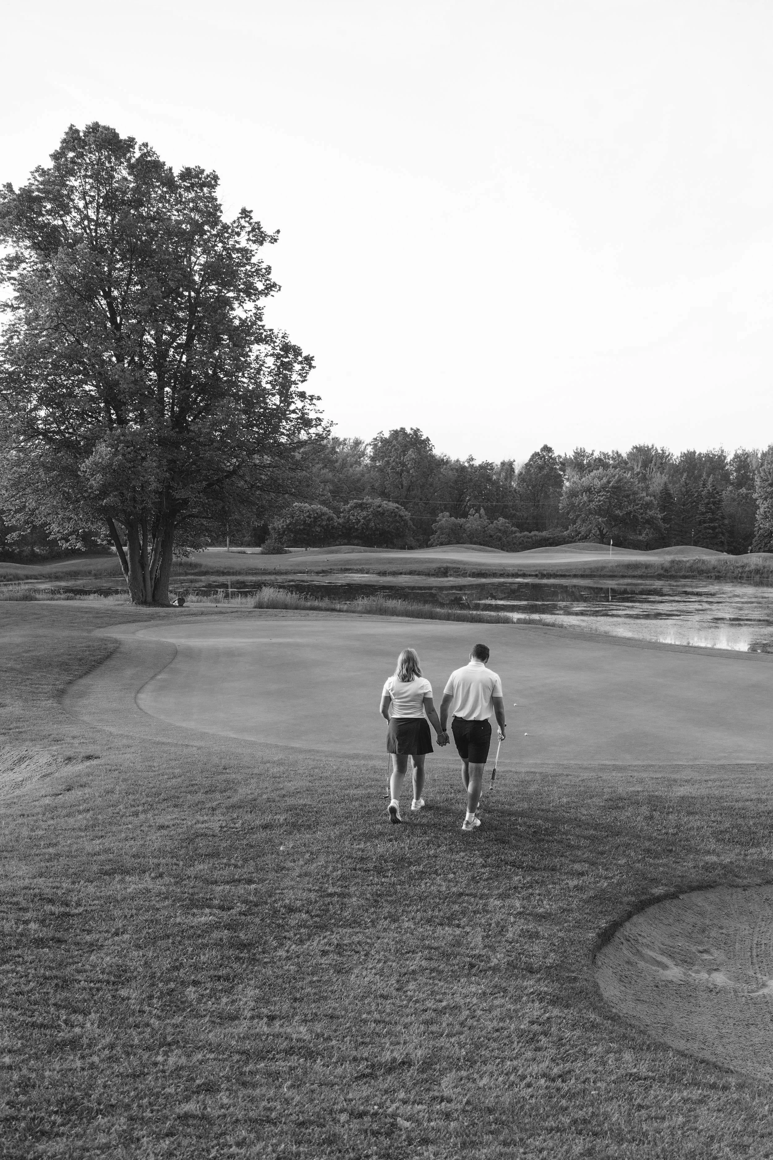 A black and white photo of a couple walking hand in hand on a golf course near a pond, with trees in the background.