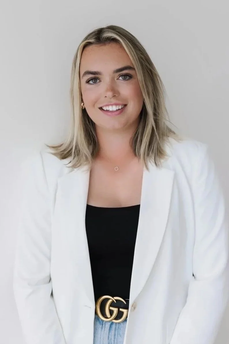 A young woman with shoulder-length blond hair, wearing a white blazer over a black top, and a gold GG belt, smiling against a plain light gray background.