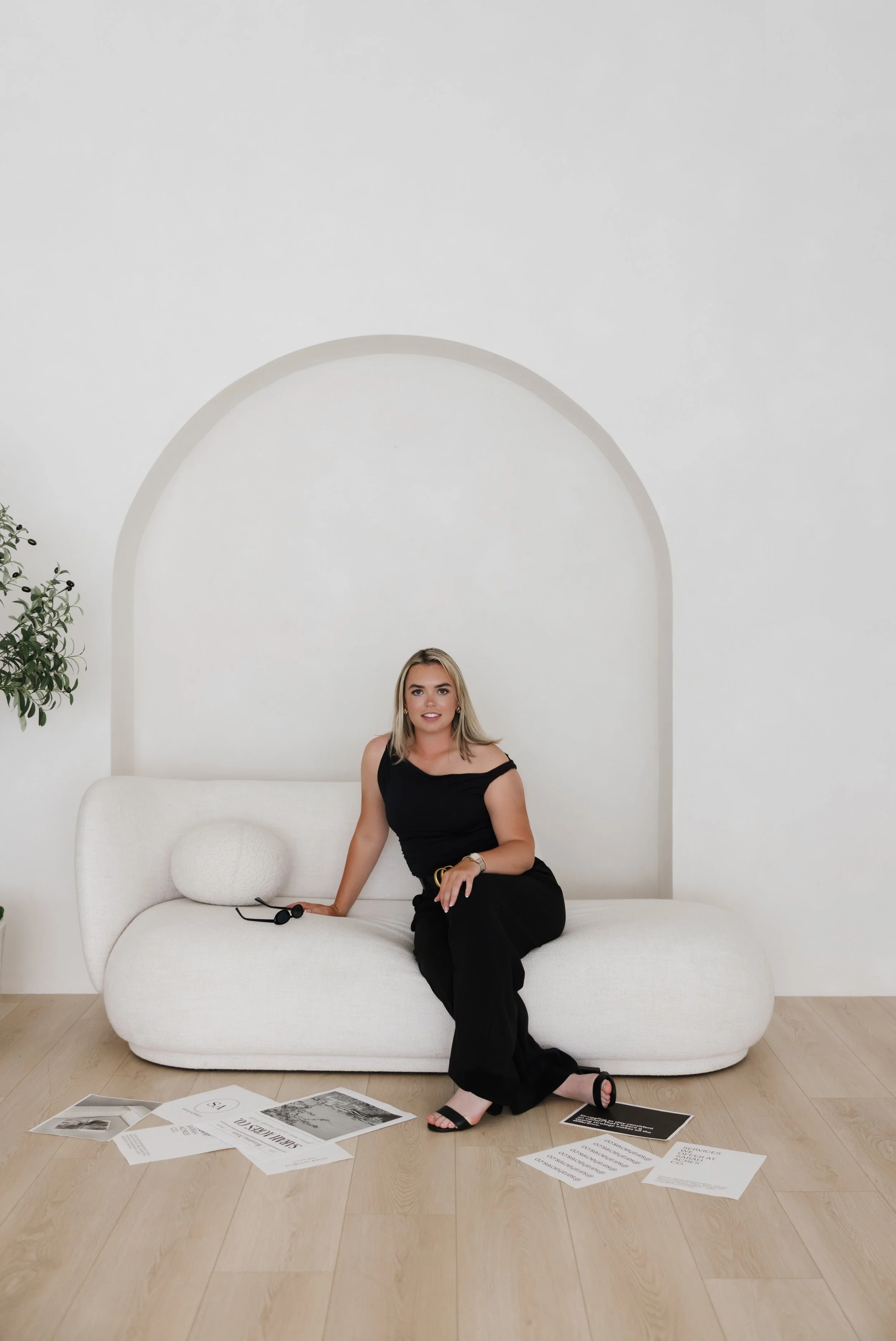 A woman in black clothes sitting on a white modern sofa with a white pillow, surrounded by scattered papers on a wooden floor, in a minimalist white room with a potted plant on the left.