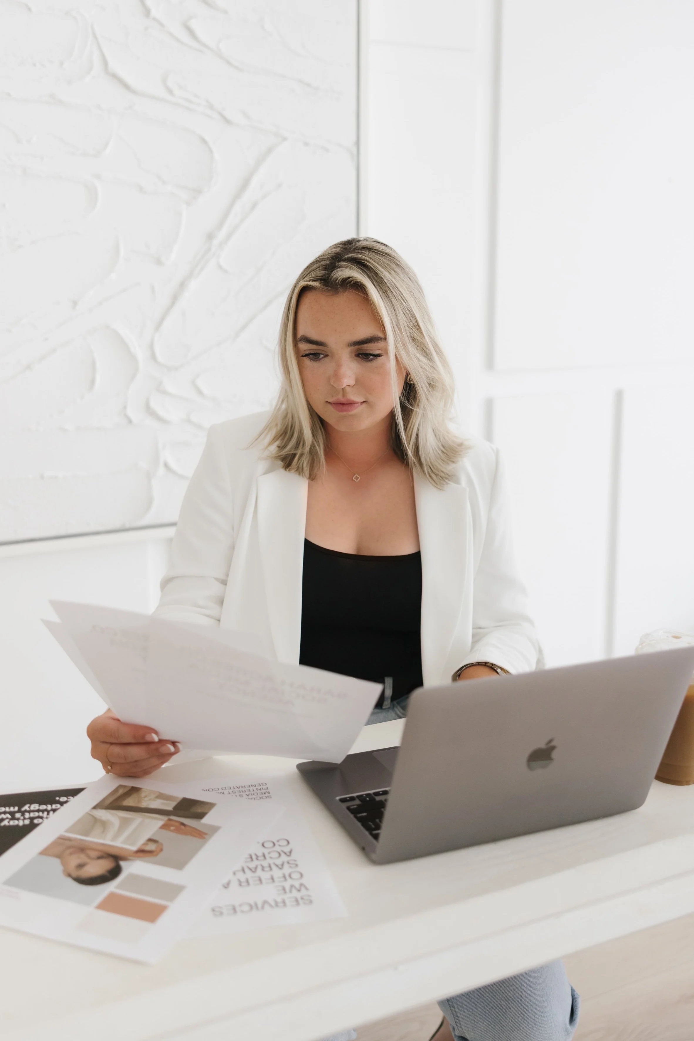 A woman with shoulder-length blonde hair working at a white desk with a laptop and papers, in a bright room with white walls.