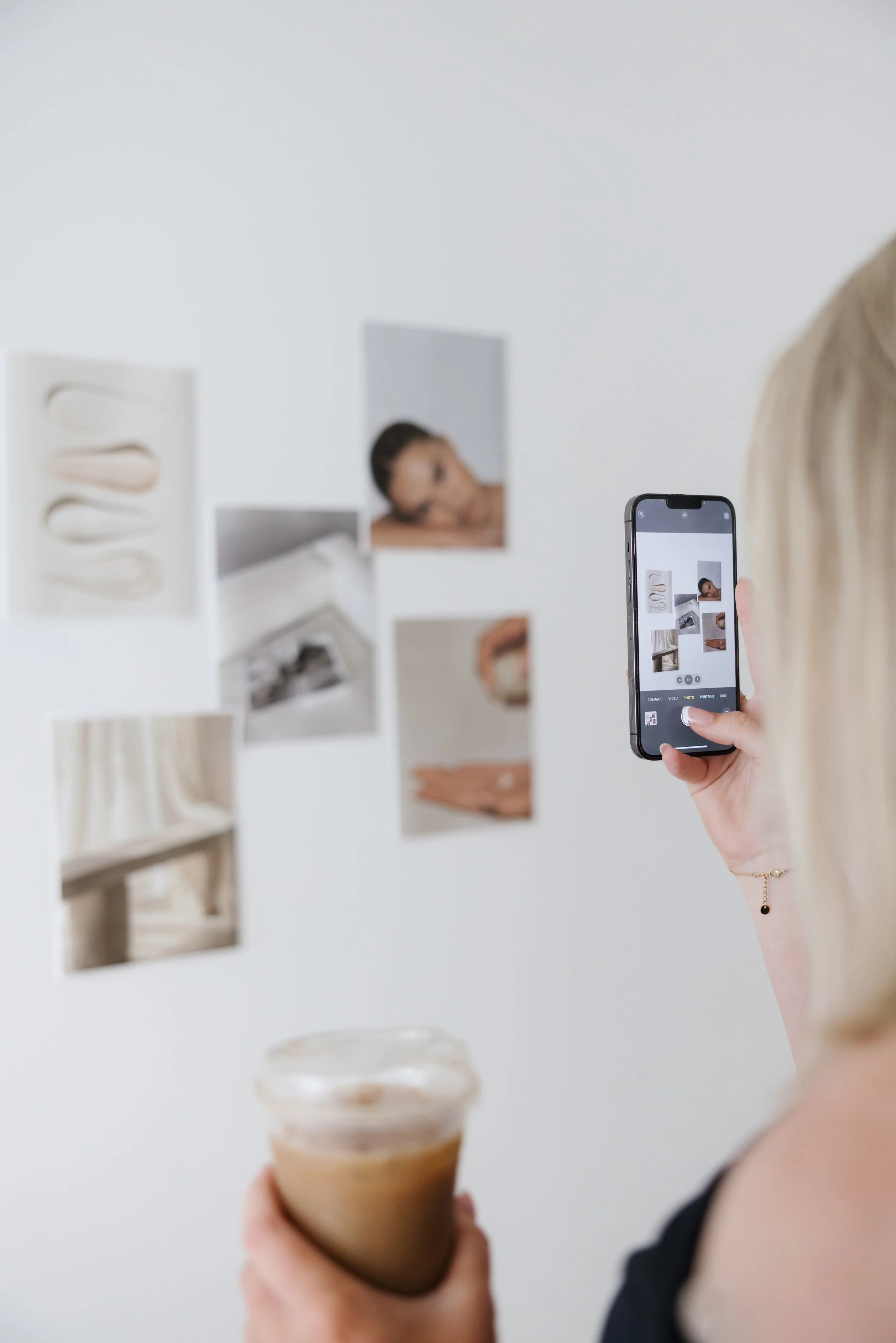 A woman with blonde hair, holding a smartphone and taking a picture of a white wall with various photos and art prints, while holding a glass of iced coffee.