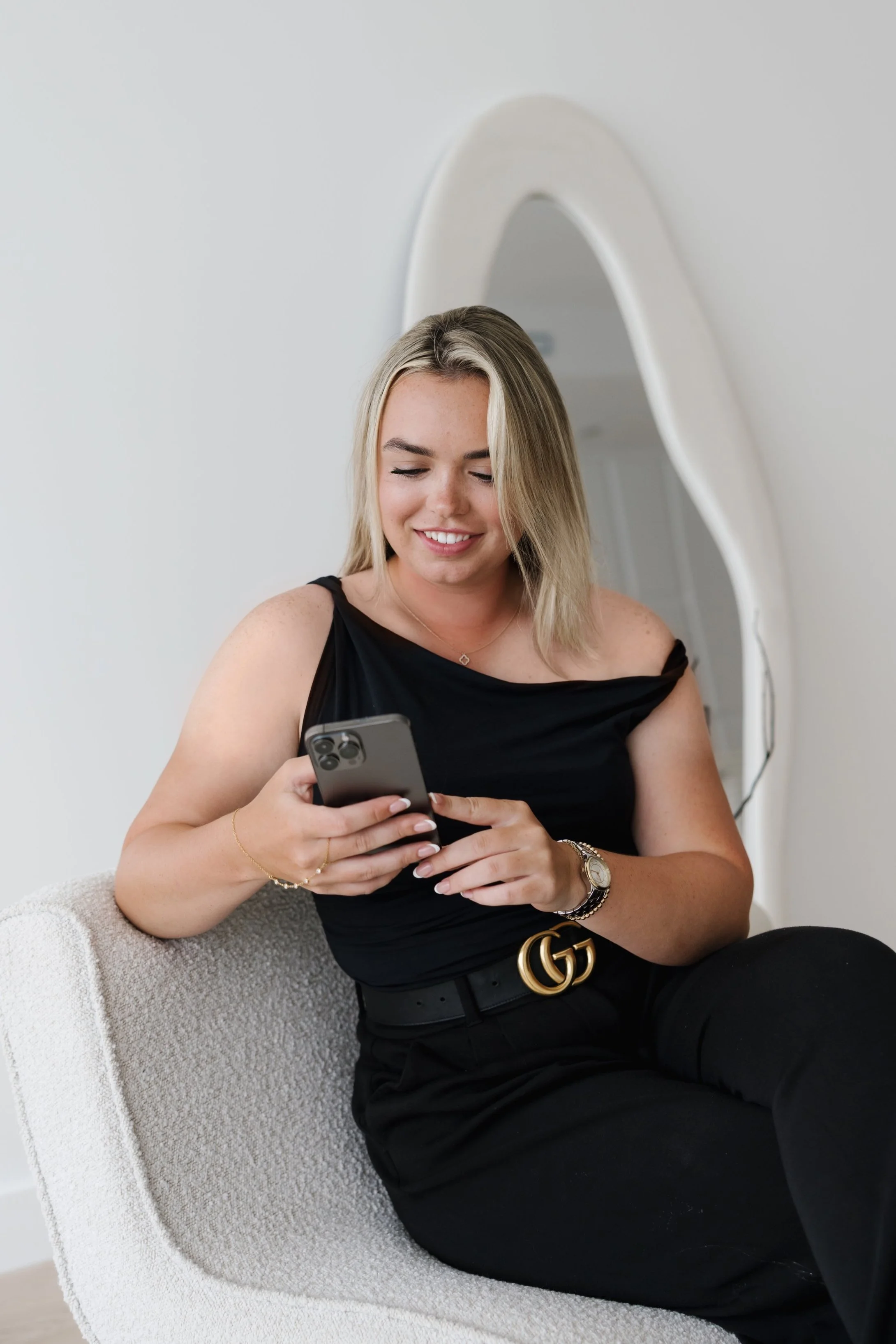 A young woman with blonde hair, smiling and looking at her phone, sitting on a white chair with a large mirror behind her