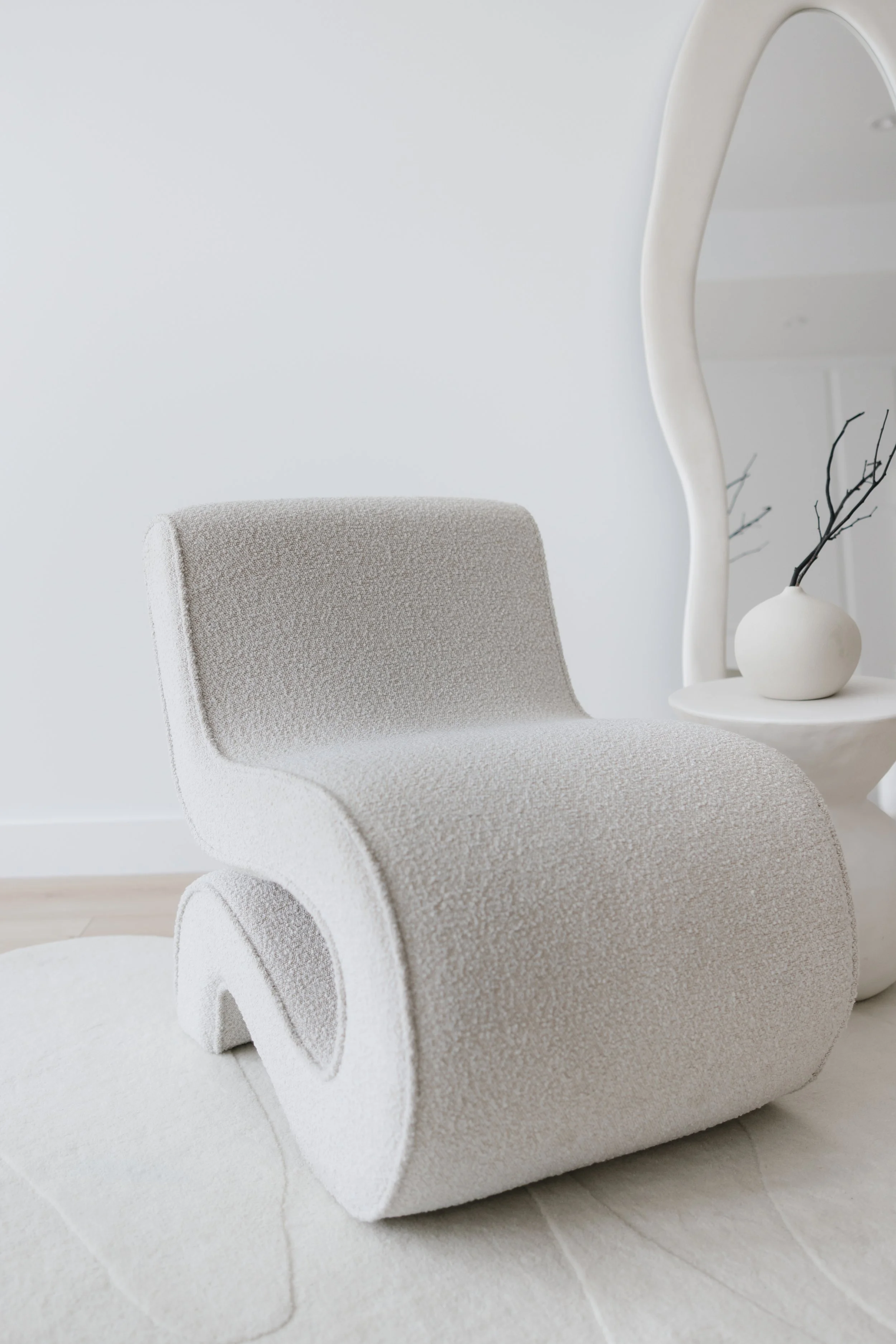 Modern white textured lounge chair with a unique curved design in a minimalist room with a white vase on a side table and a large mirror.