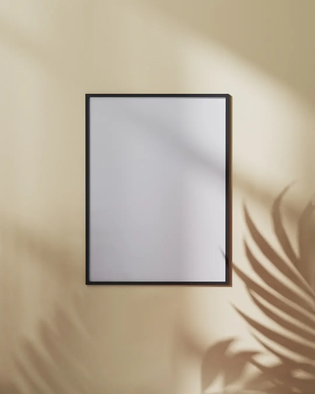 A rectangular mirror with a black frame hanging on a beige wall, with a shadow of a leaf on the wall to the right.