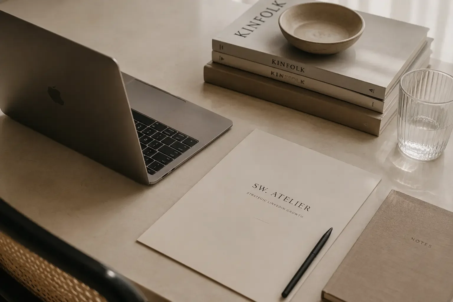Et skrivebord med en åben MacBook, en bunke bøger, en tom skål, et glas vand, en notesbog, et dokument med overskriften 'SW: ATELIER', LinkedIn Optimering