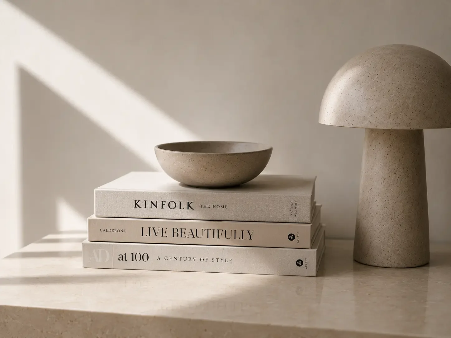 Et bord med tre bøger, en lille skål på toppen, og en mushroom-lignende bordlampe i beige, med naturlige farvetoner og simpel, minimalistisk stil.