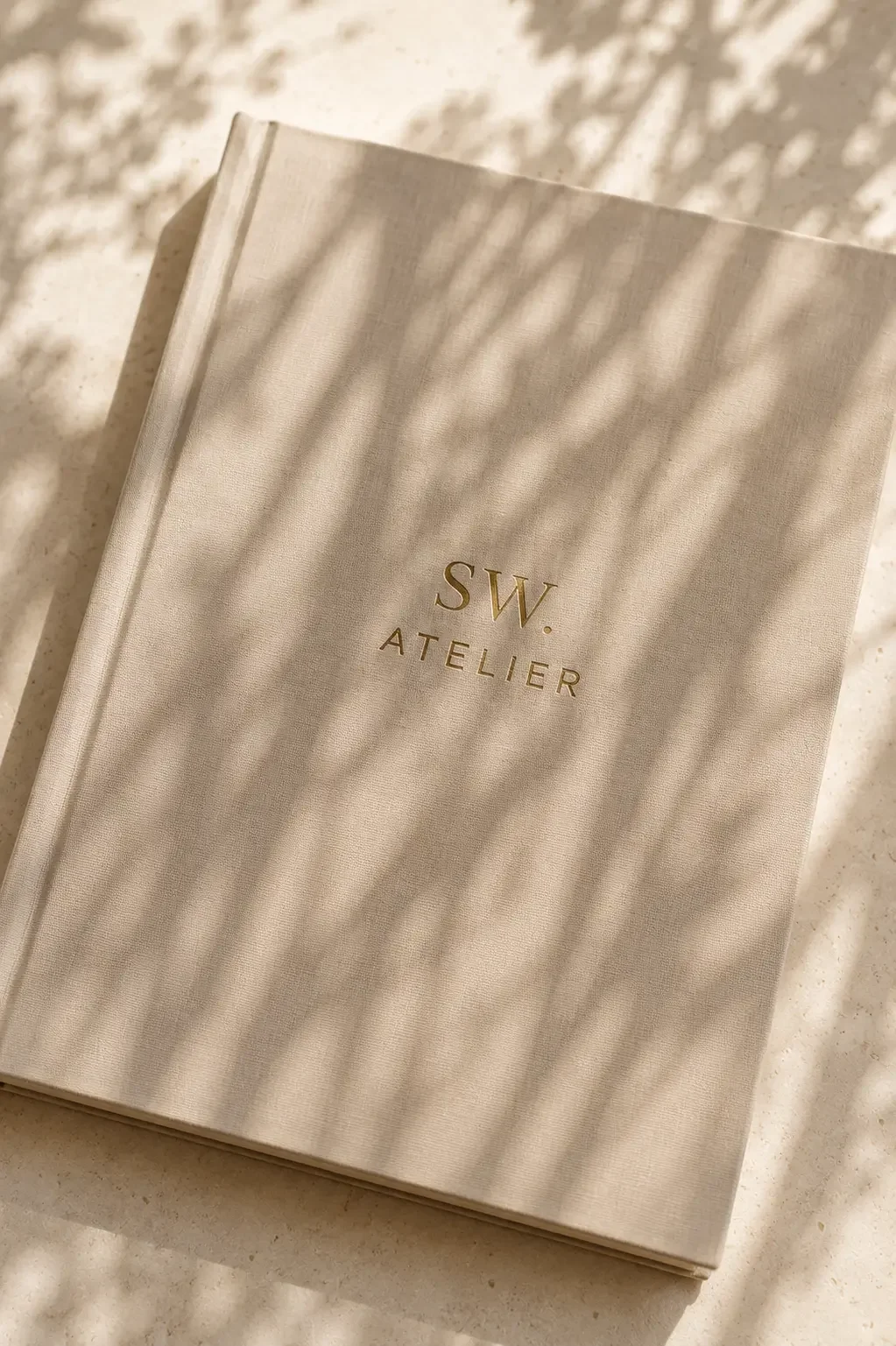 En beige bog med teksten 'SW. ATELIER' i guld på forsiden, liggende på en lys overflade med skygger af blade kastet ovenfra.
