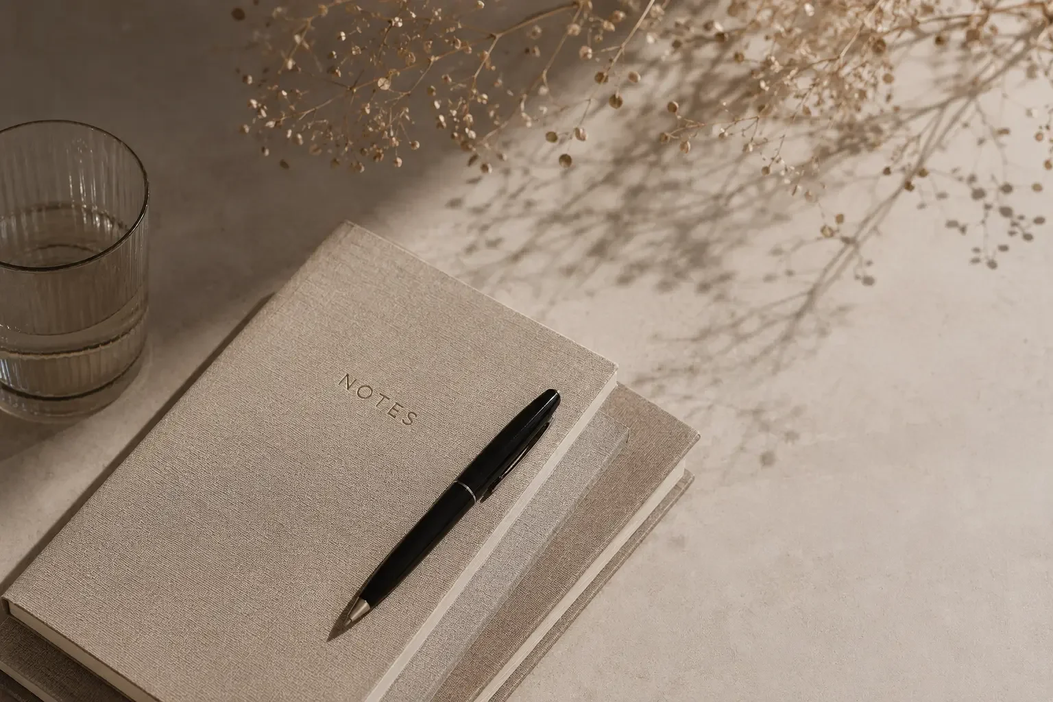 En beige notesbog med teksten 'NOTES' på forsiden, en sort pen, et krystallglas med vand, og tørrede blomster, der kaster skygger på bordet.